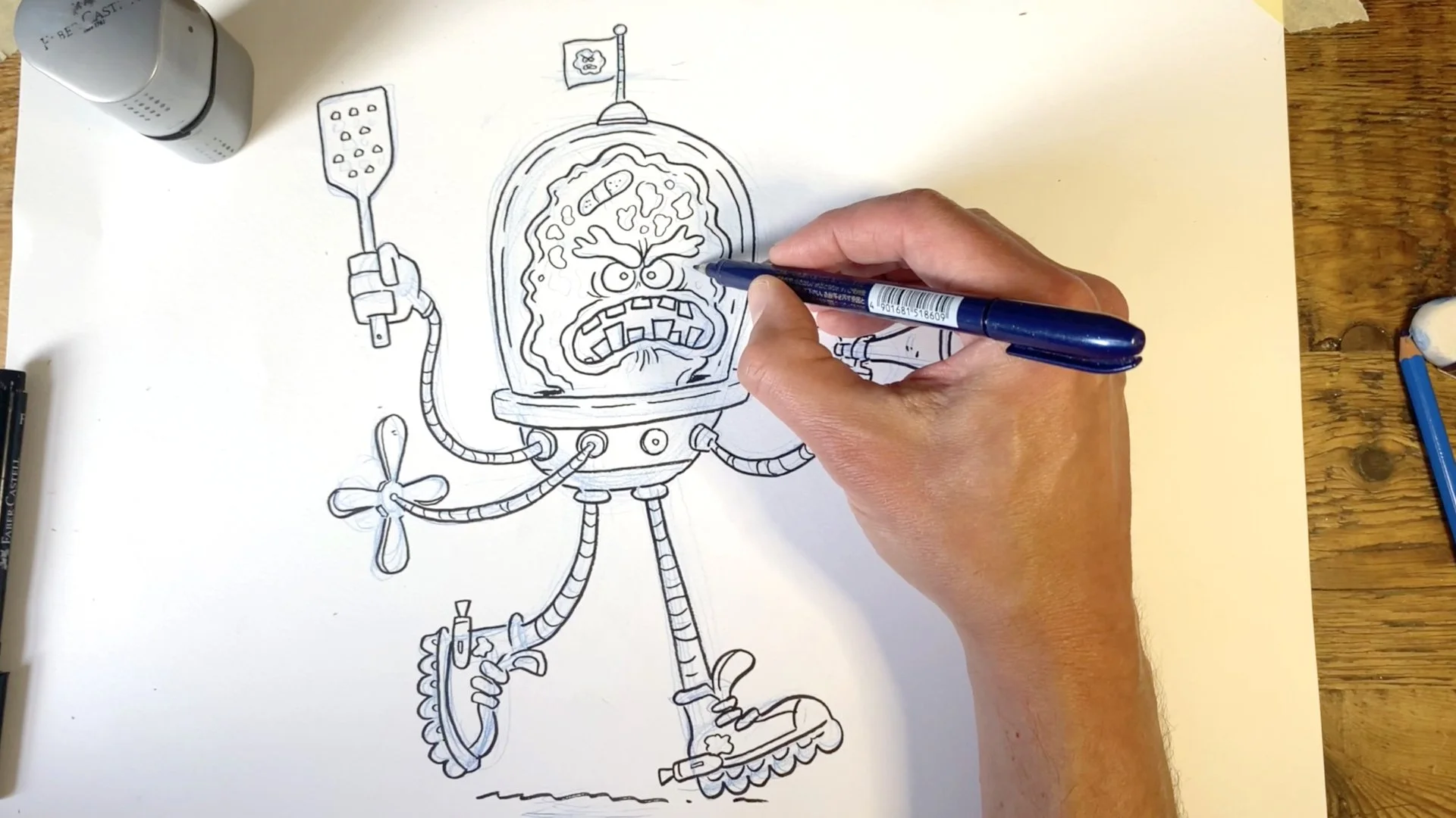Hand drawing a cartoon monster with a helmet and propeller on paper, surrounded by pens and drawing tools on a wooden table.