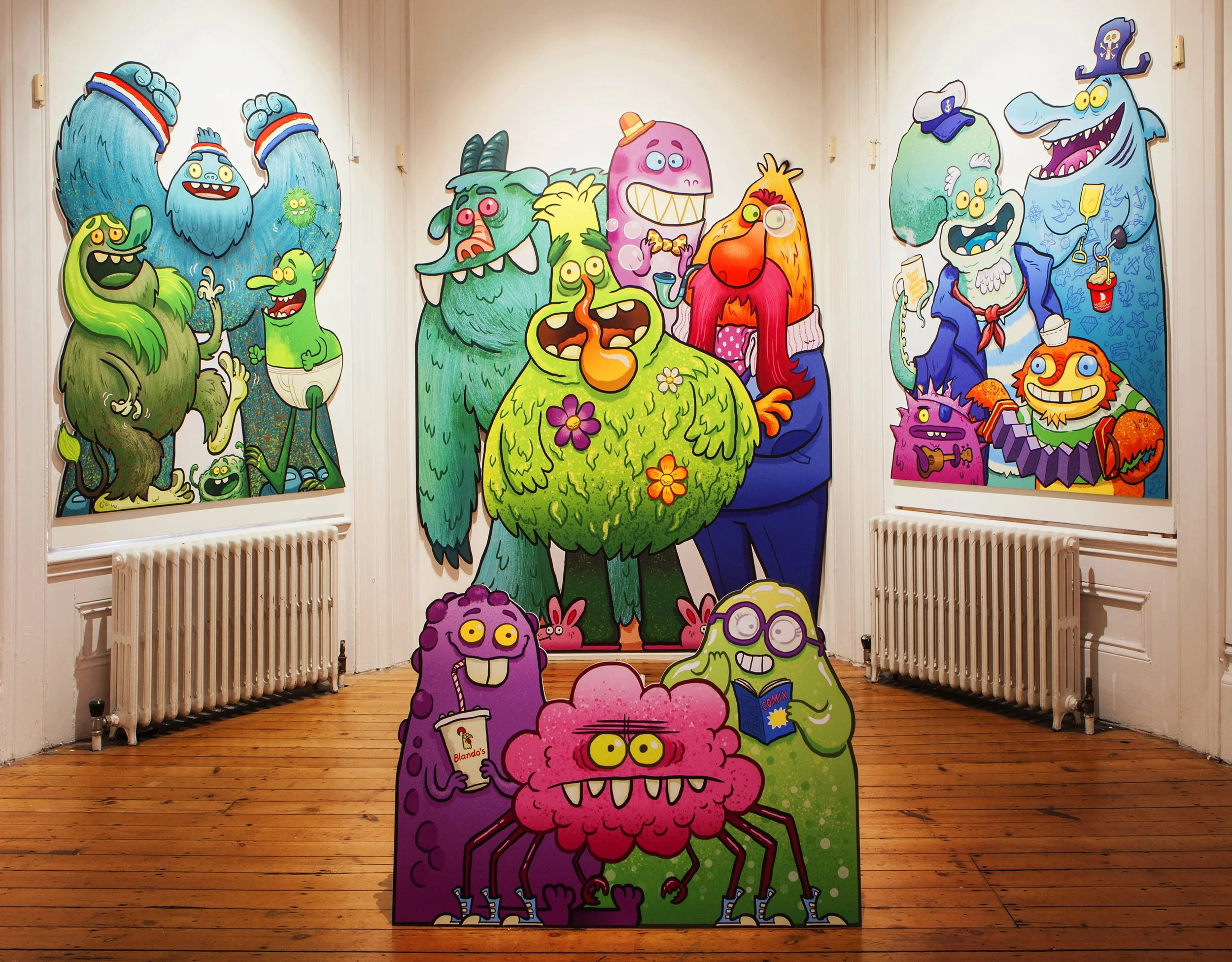 Colorful cartoon monster cutouts on a gallery wall, featuring varied designs, colors, and expressions, with some holding objects like books and drinks.
