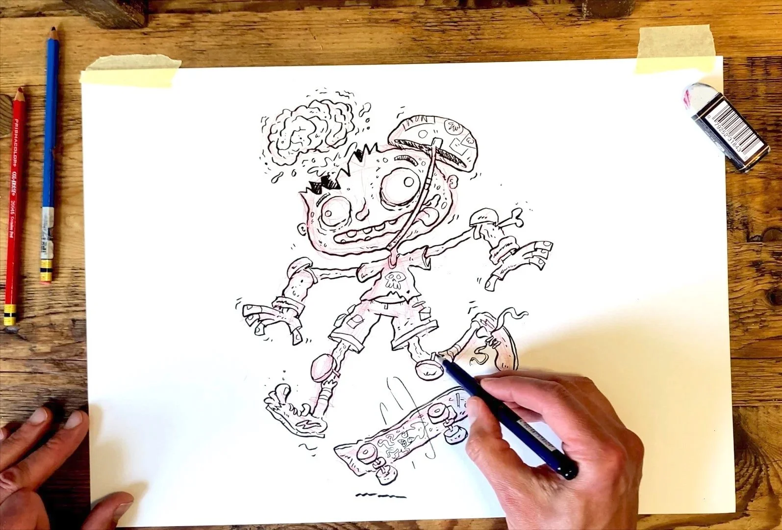 A person is drawing a cartoon character of a zombie skateboarder with a skateboard and an umbrella, on a piece of white paper on a wooden table. The character has a large head, wide eyes, and a tattered outfit with a skull symbol on the shirt.
