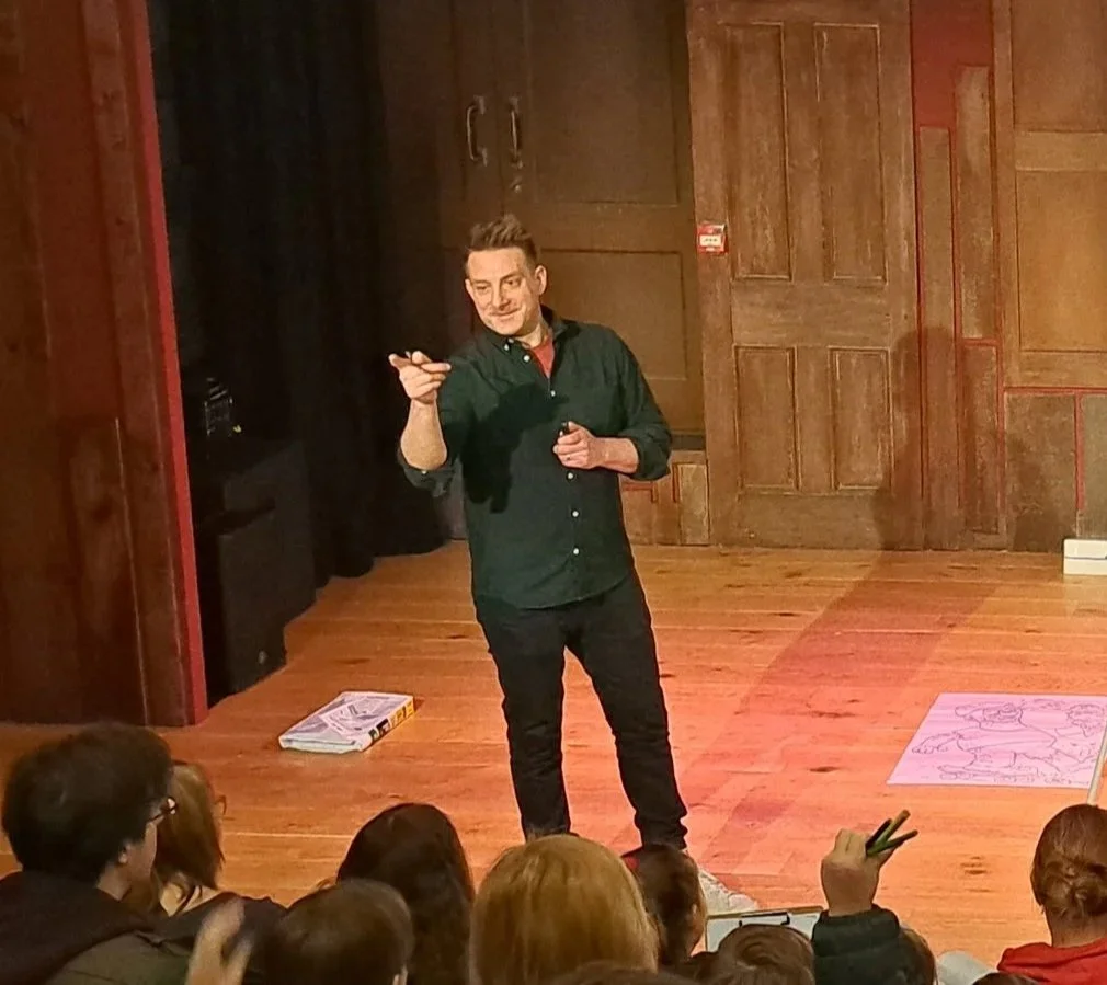 A man is standing on stage in front of an audience, speaking or performing. The stage has wooden floors and wooden paneling in the background. There are several people sitting and some raising their hands in the audience.