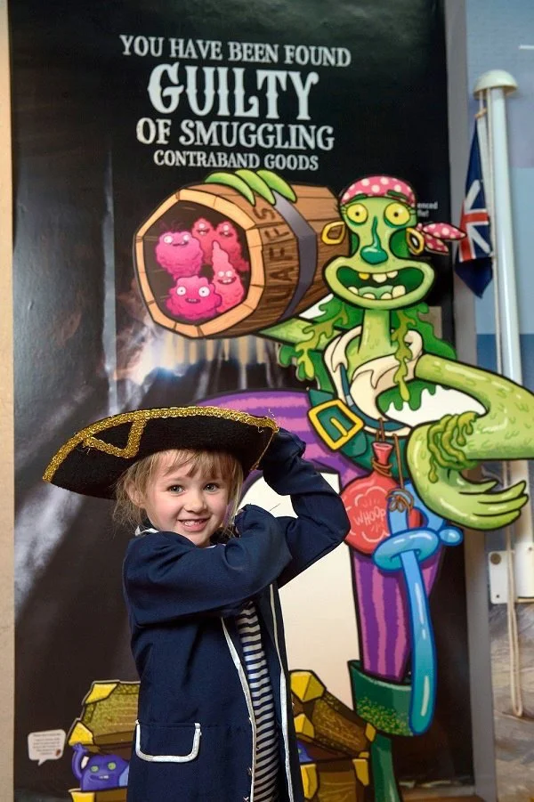 A young girl wearing a pirate hat, standing in front of a pirate-themed poster with a cartoon pirate and a barrel labeled "ALERT" containing pink smuggling critters. The girl is smiling and holding her pirate hat.