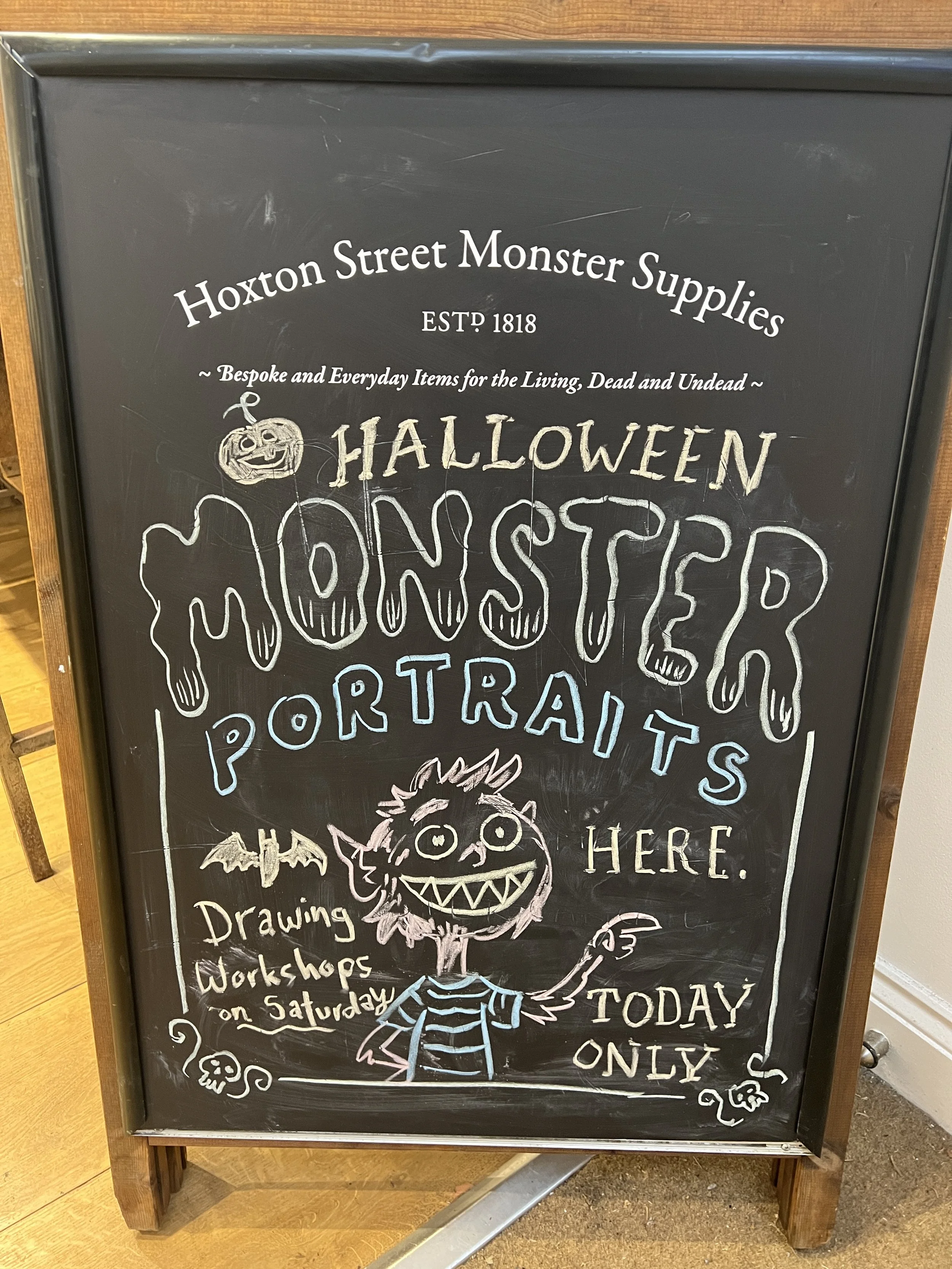 A chalkboard sign advertises Halloween-themed items and workshops at Hoxton Street Monster Supplies, established in 1818. The sign includes drawings of a smiling ghost, a cartoonish monster with sharp teeth, a bat, a skull, and a Frankenstein-style figure.