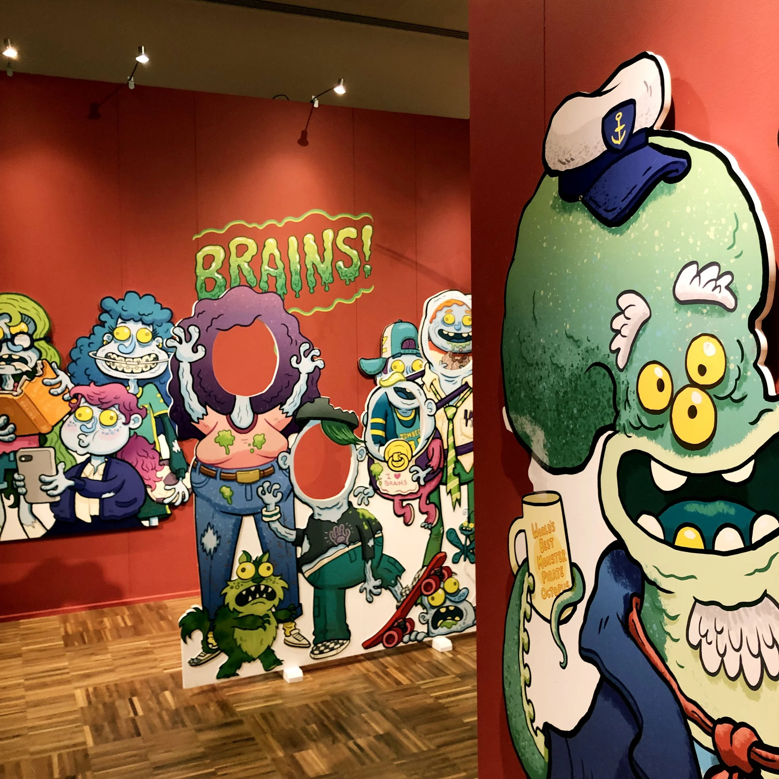 Colorful cartoon illustrations of various zombies and monsters displayed as standees in an art exhibit, with the word 'BRAINS!' in bright green on a red background.