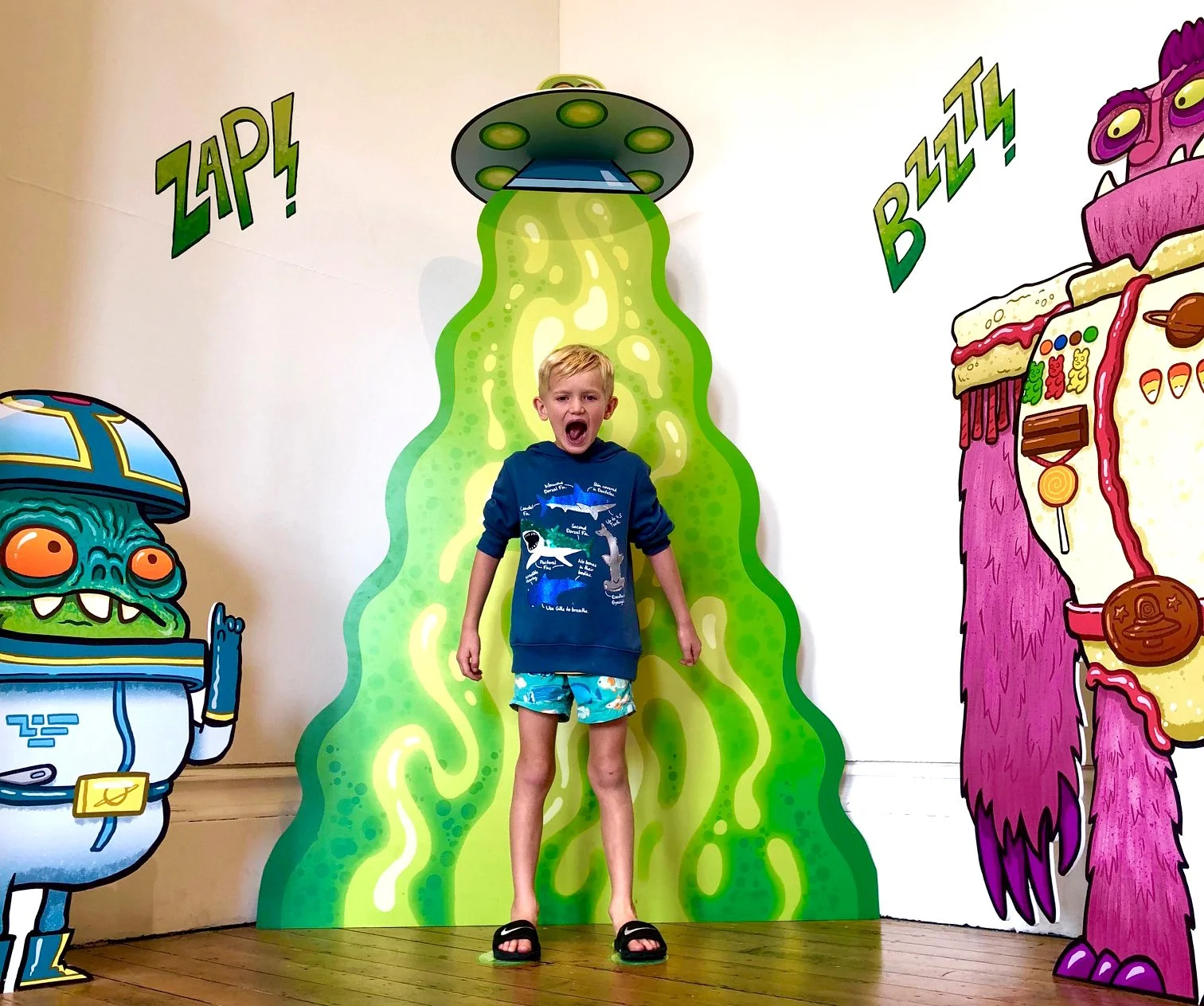 A young boy standing in front of colorful cartoon alien and monster cutouts, with a green and yellow slime-like background. The boy is wearing a blue hoodie with marine animal illustrations, blue shorts, and black sandals, and has his mouth open in an excited expression.