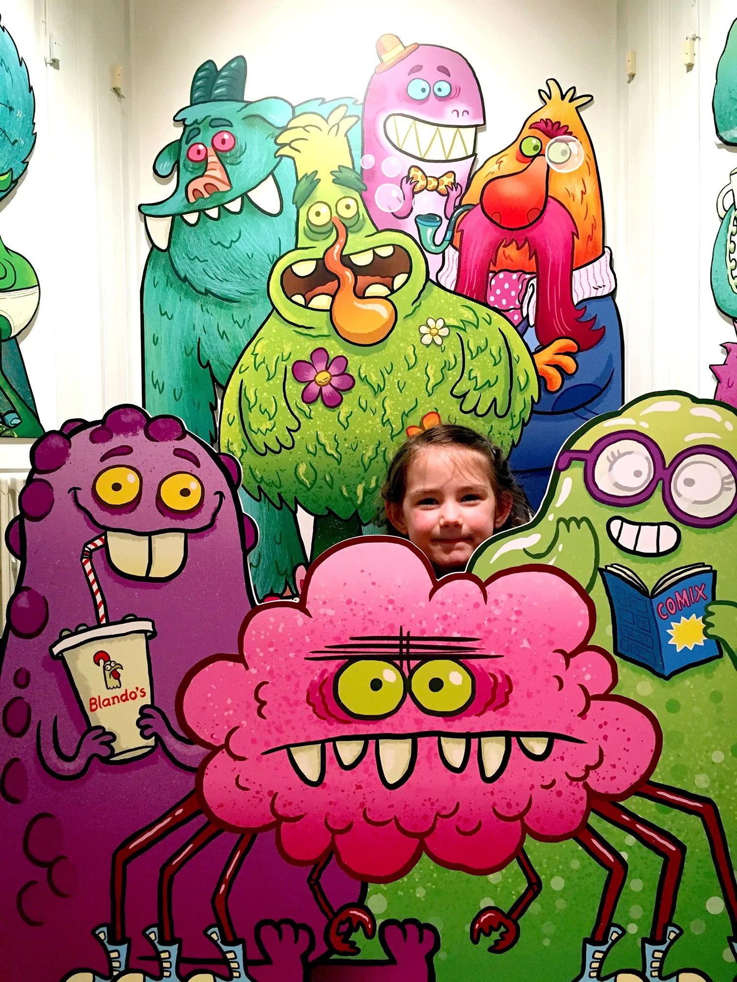 Colorful cartoon monsters share a fun scene, some holding treats, drinks, and a comic book, set against a vibrant, illustrated background.