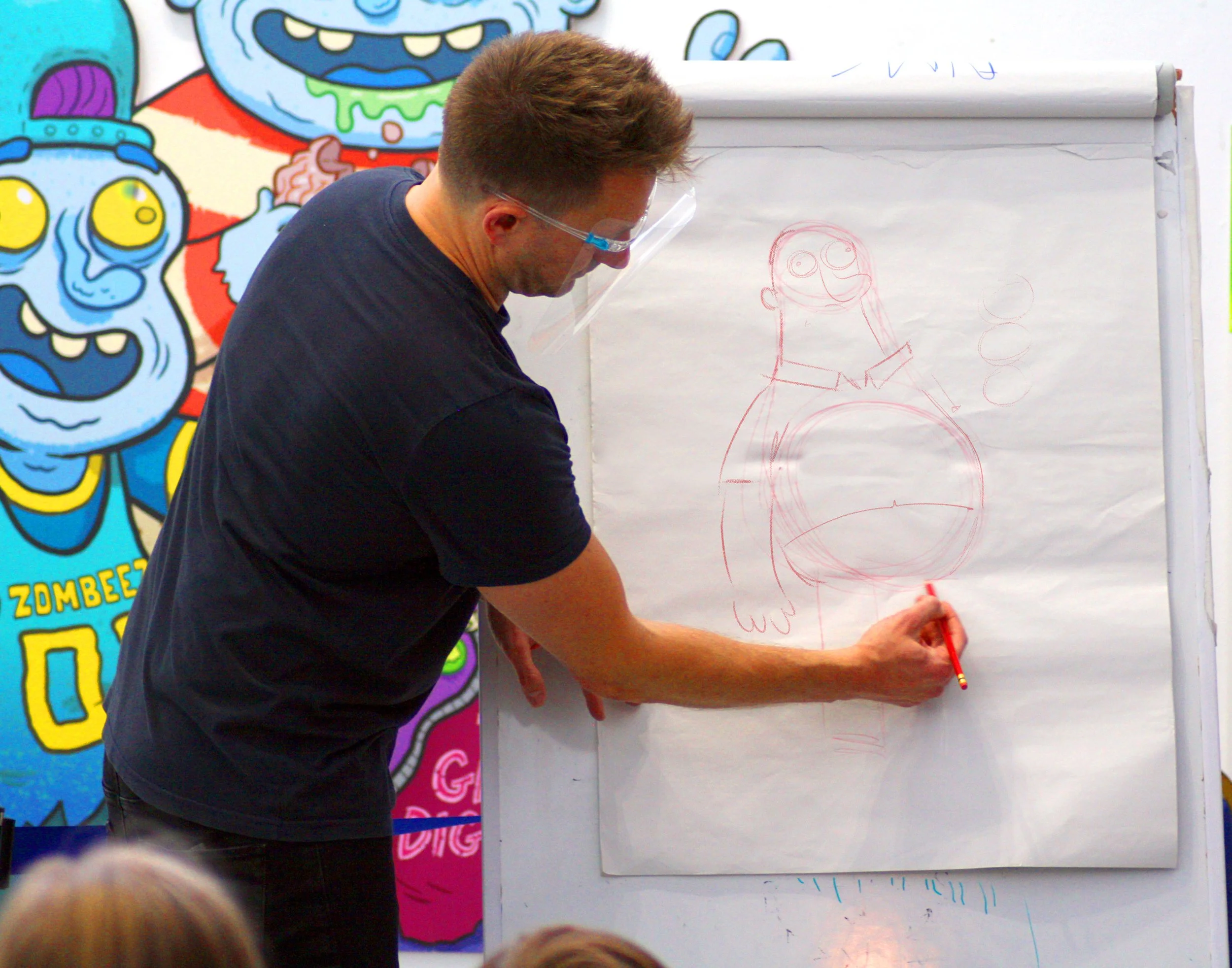 A man drawing a cartoon character on a whiteboard with a red marker, in front of a colorful graffiti-style mural.