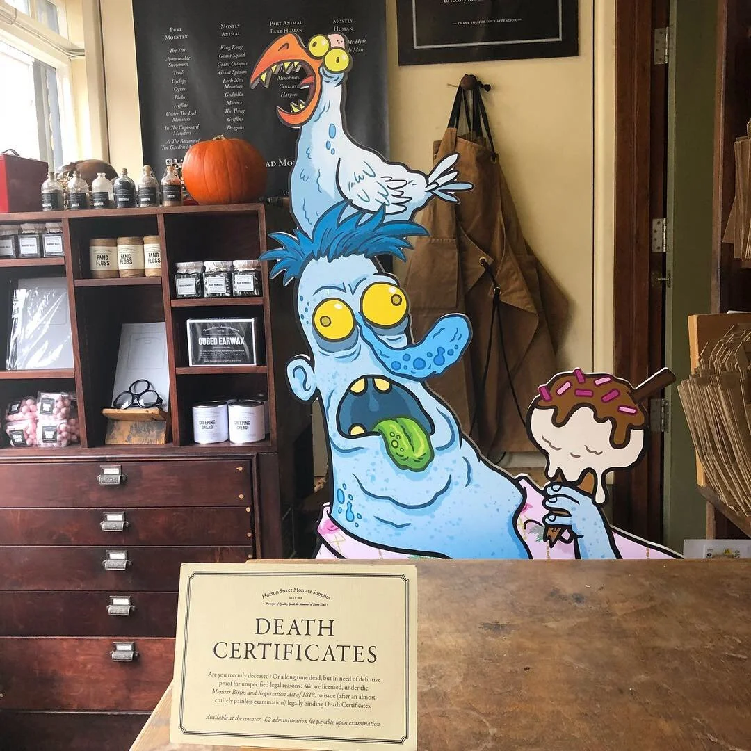 Colorful cartoon figure of a zombie with blue skin, yellow eyes, a green tongue, and blue hair, holding an ice cream cone with pink sprinkles and chocolate sauce, with a cartoon bird on top of its head and an orange pumpkin on the shelf behind.