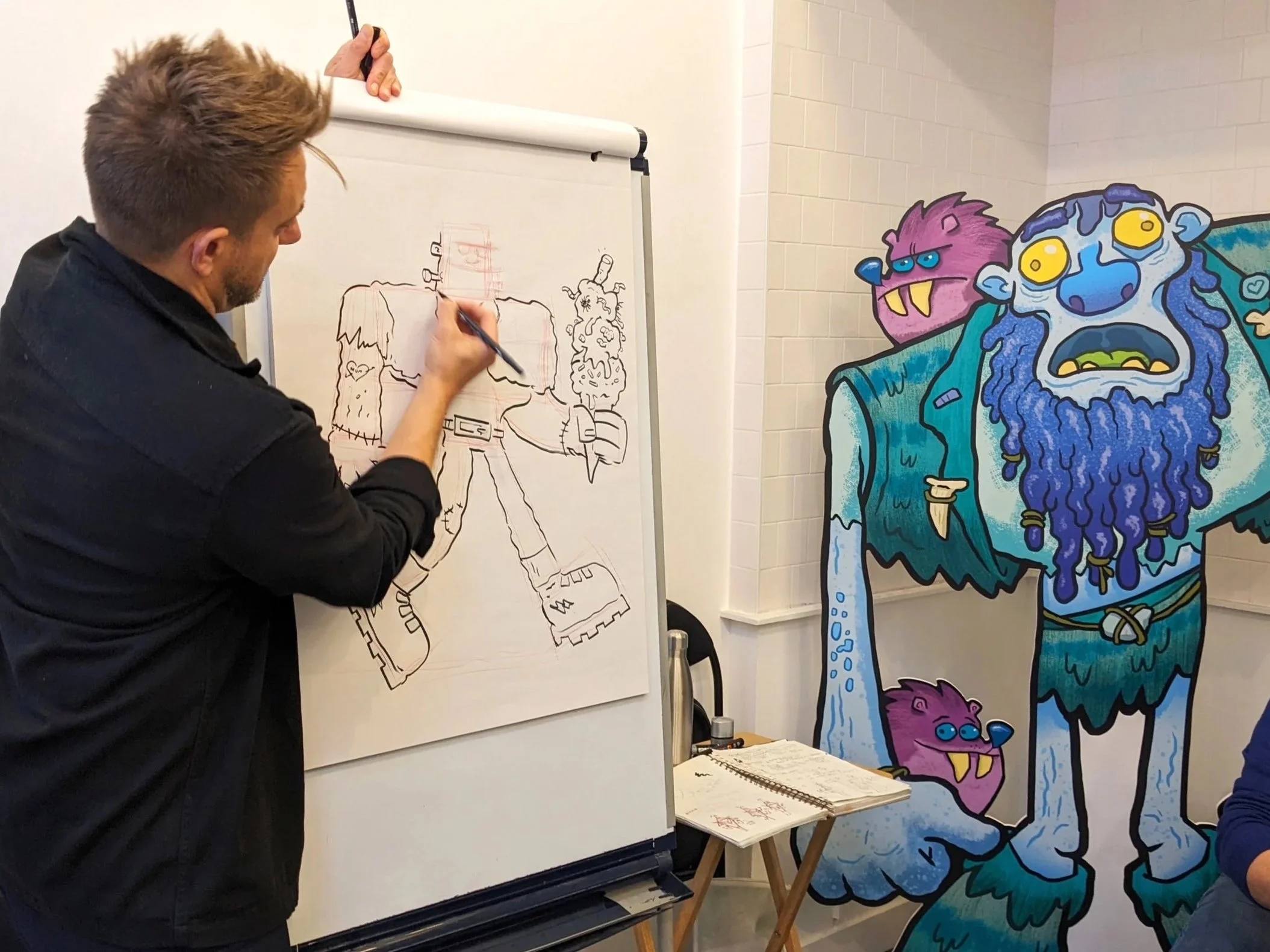 A man drawing a character on a whiteboard, with colorful cartoon monsters standing to the right