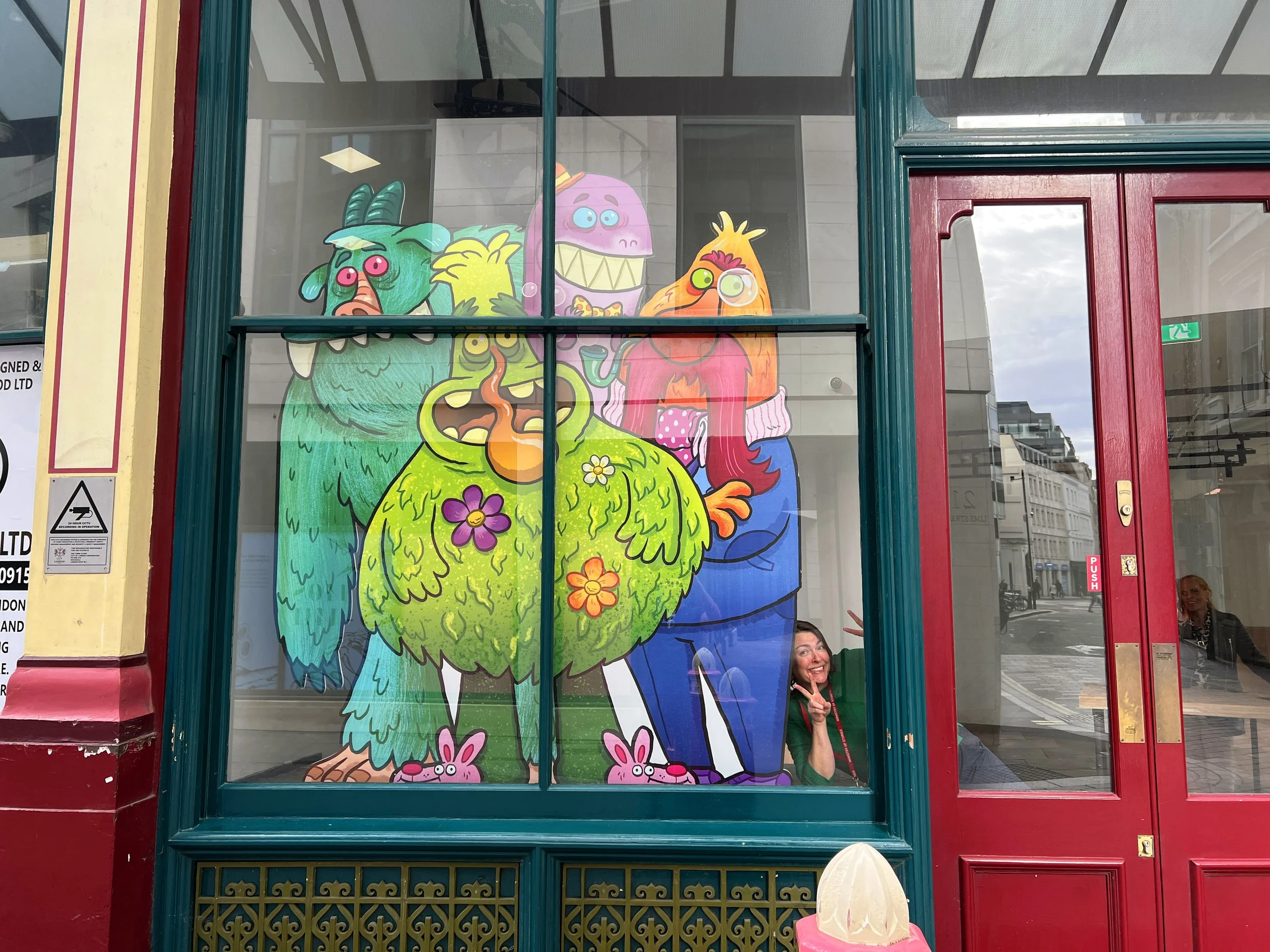 Colorful cartoon monster characters painted on a glass storefront window, with a woman in a green jacket and red lanyard standing outside making a peace sign.