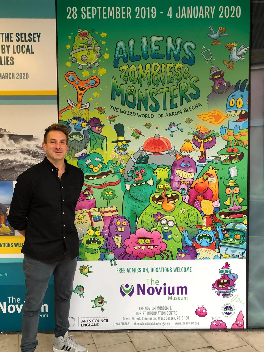 A man standing in front of a colorful poster for an exhibit titled 'Aliens, Zombies & Monsters' at The Novium Museum. The poster features cartoonish illustrations of various monsters, aliens, and zombies in vibrant colors, with the exhibit running from September 28, 2019 to January 4, 2020.