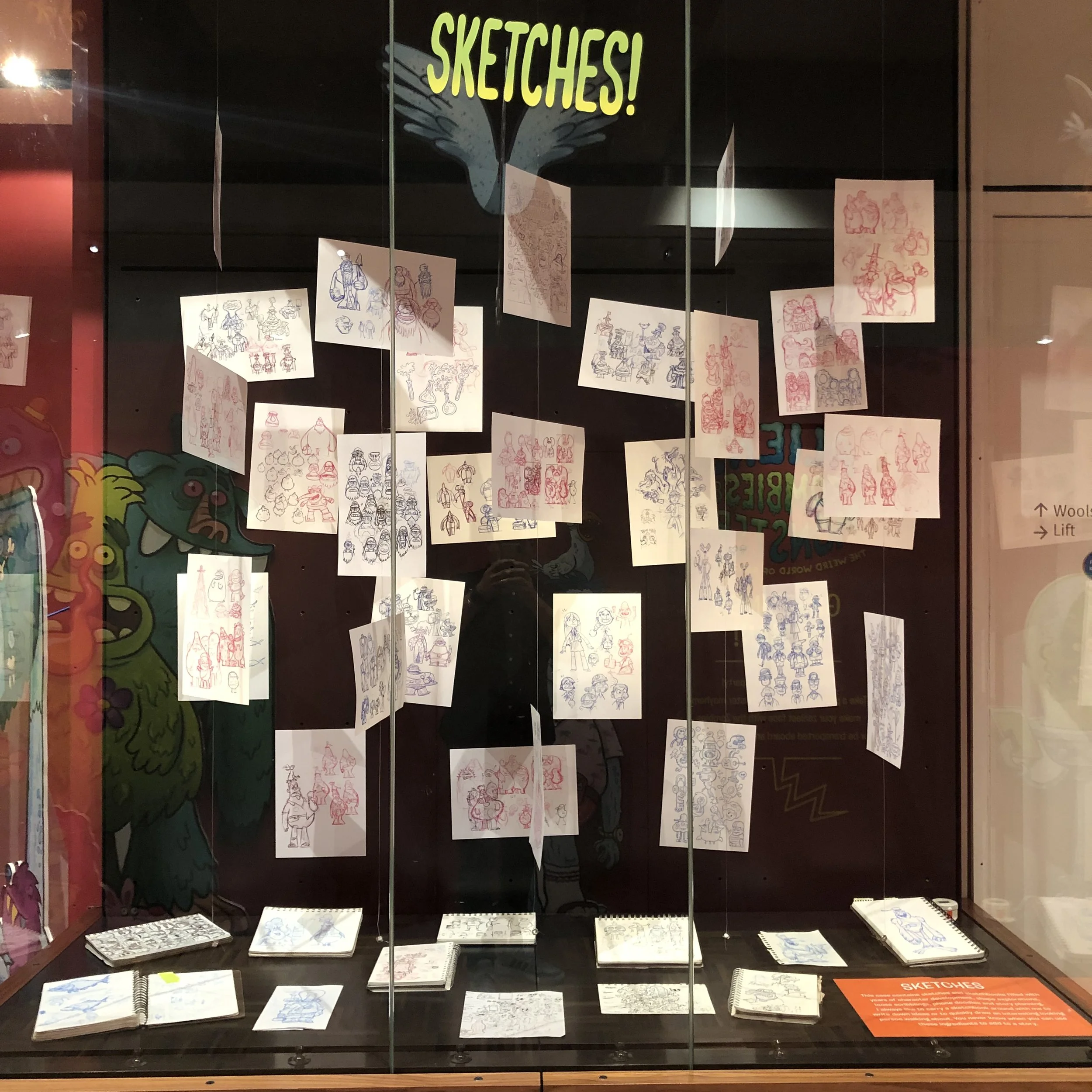 Display of sketches and drawings of people and characters, hanging in a glass case with a banner that says 'SKETCHES!' at the top.