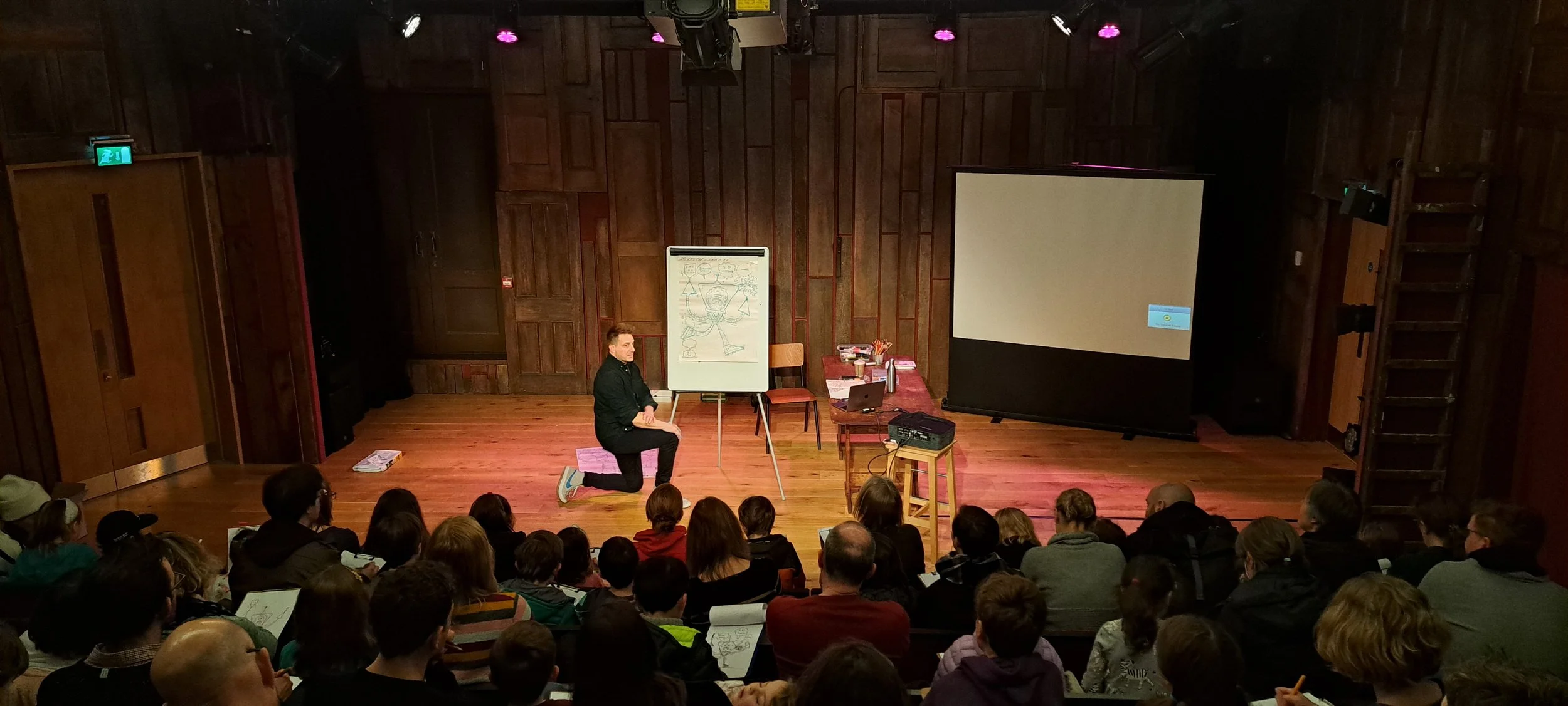 A man kneeling on one knee on a stage giving a presentation to a large audience. There is a whiteboard with a caricature drawing, a table with a laptop and books, and a large projection screen.