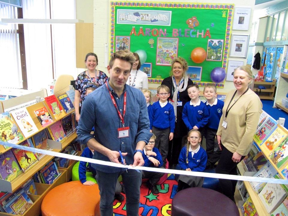A man is cutting a ribbon during a children's book event at a library, with children and adults smiling behind him, bookshelves on both sides, and colorful decorations on the bulletin board in the background.