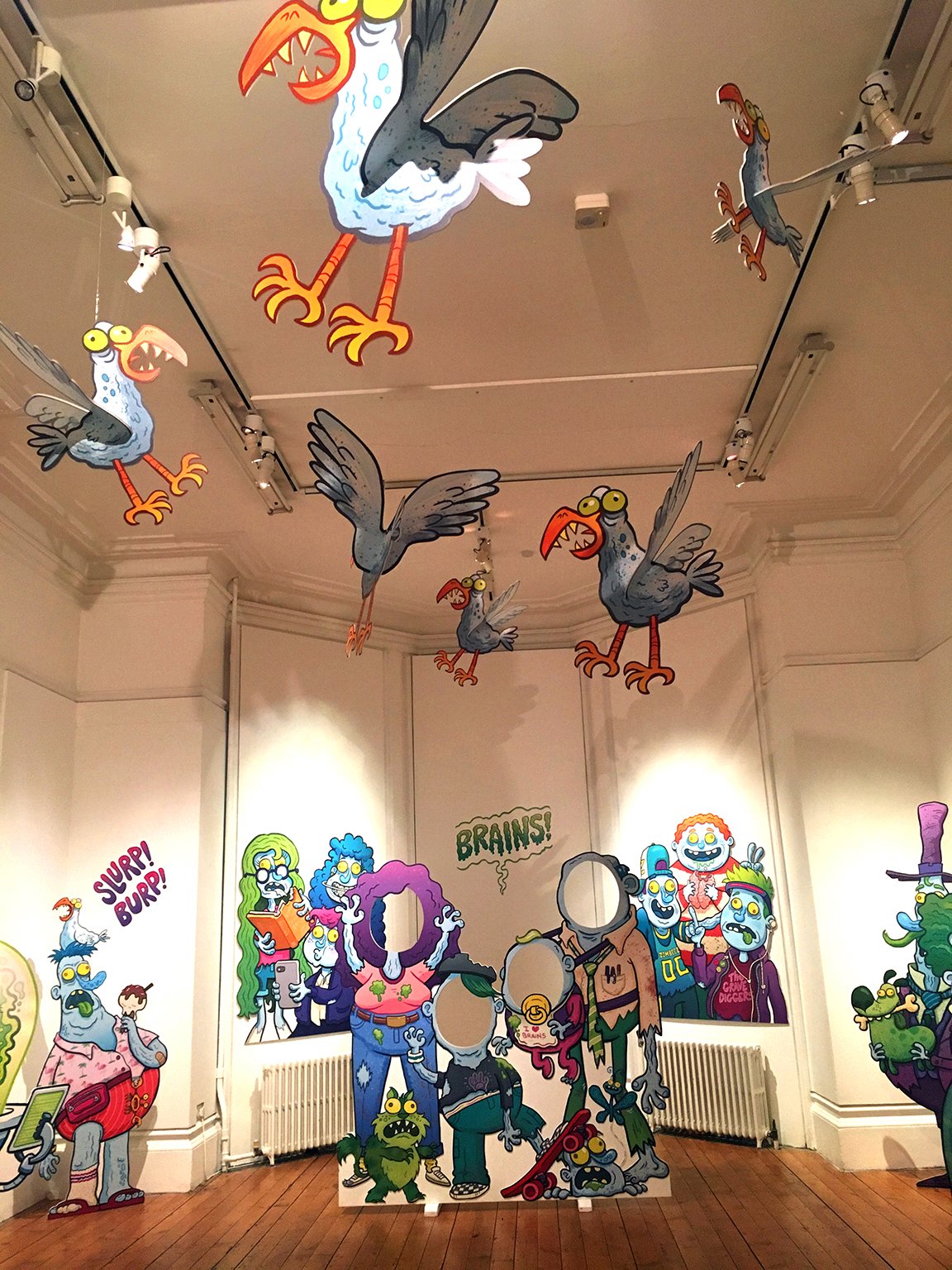 Colorful cartoon-style illustrations of zombies and birds with exaggerated features and bright colors, set in a room with a wooden floor and white walls, featuring signs with words like 'BRAIN!' and 'SLURP! BURP!'.