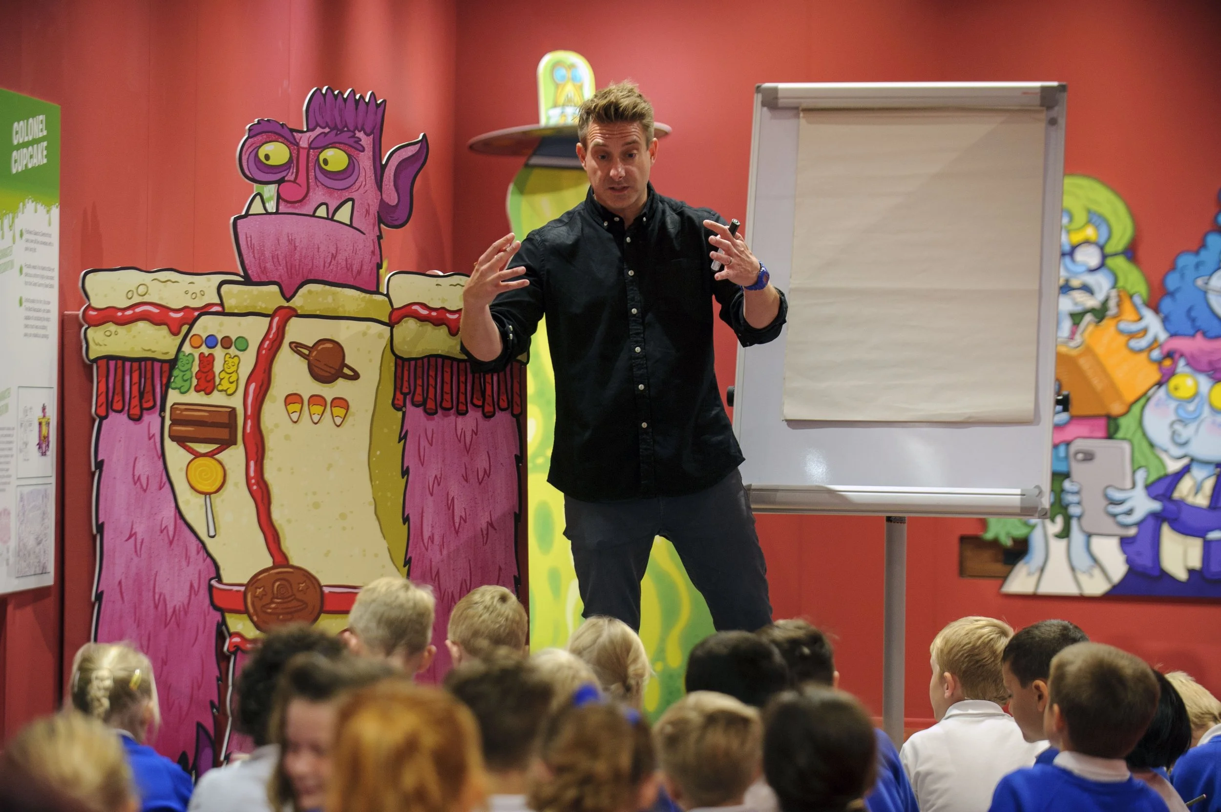 A man giving a presentation to a group of children in a colorful, cartoon-themed room. The background features large cartoon character cutouts and a whiteboard.