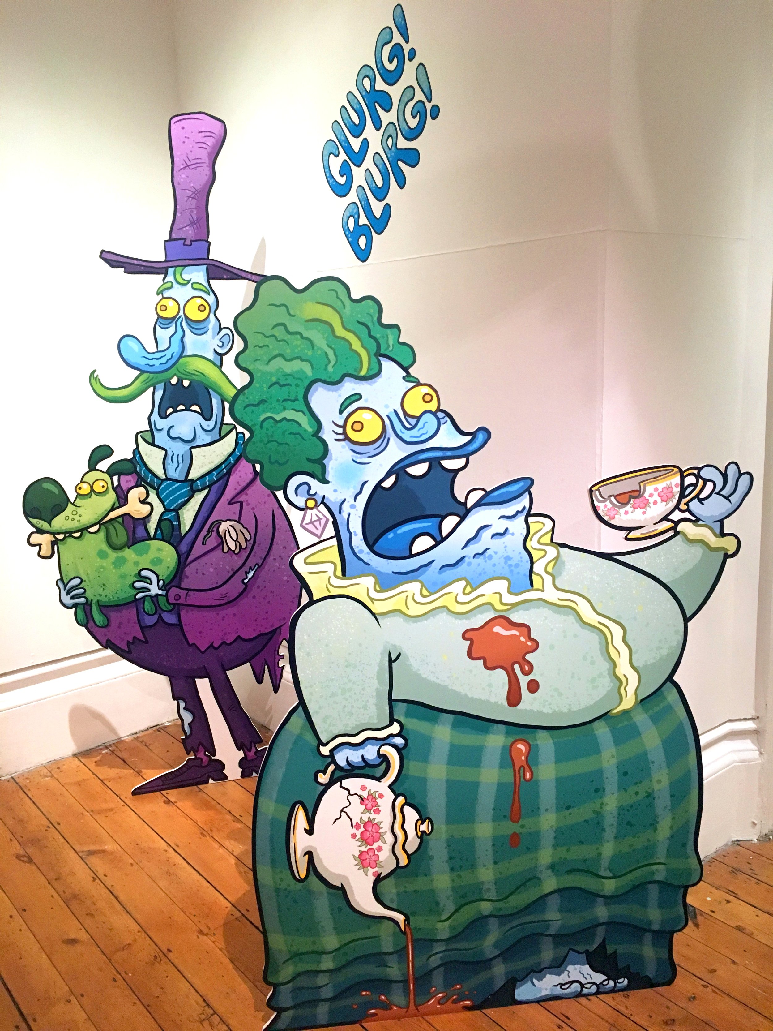 Colorful cartoon illustration of a group of ghoulish characters with exaggerated features, including one with green hair holding a tea cup, another with a long mustache and purple suit, and a small green creature. The characters appear to be in a spooky, humorous scene with the words 'GHOULS! BLOO! G!' written in blue on the wall.