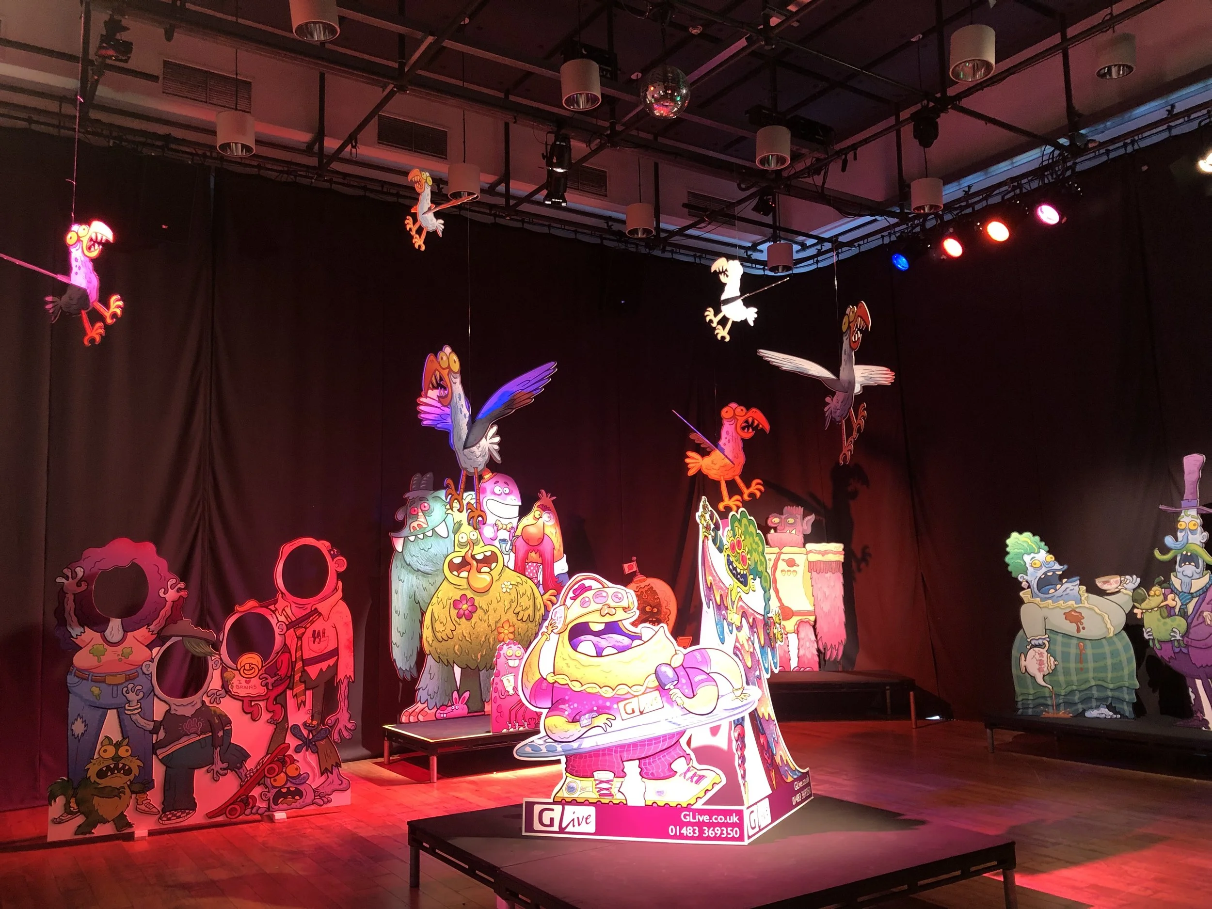 Colorful, illuminated cardboard cutouts of various cartoonish monsters and creatures displayed on a stage with a black backdrop, under stage lights in an indoor venue.
