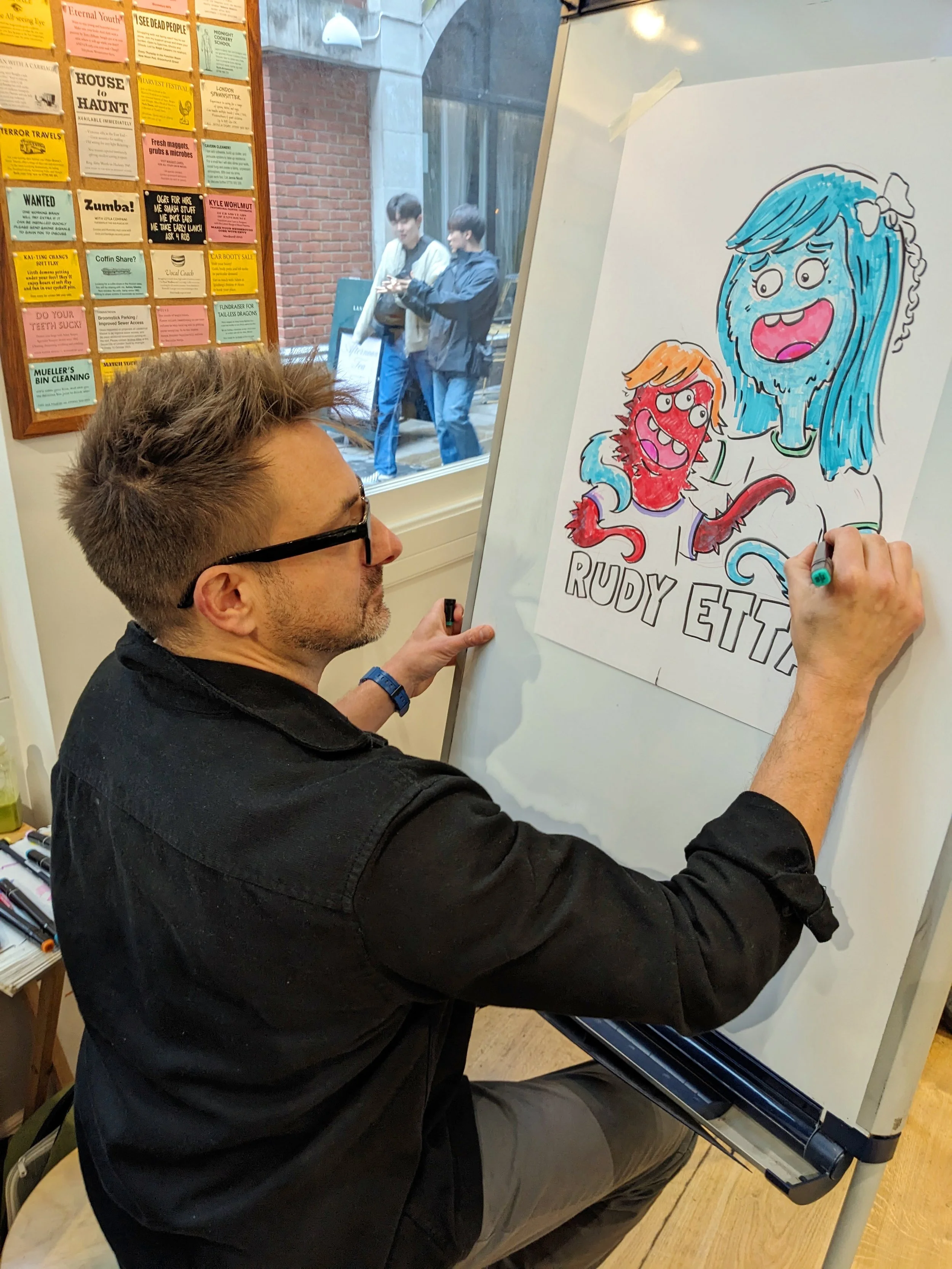 A man with glasses seated at a piano, drawing a colorful cartoon poster of two girls with exaggerated facial expressions, with a window and two young men outside in the background.