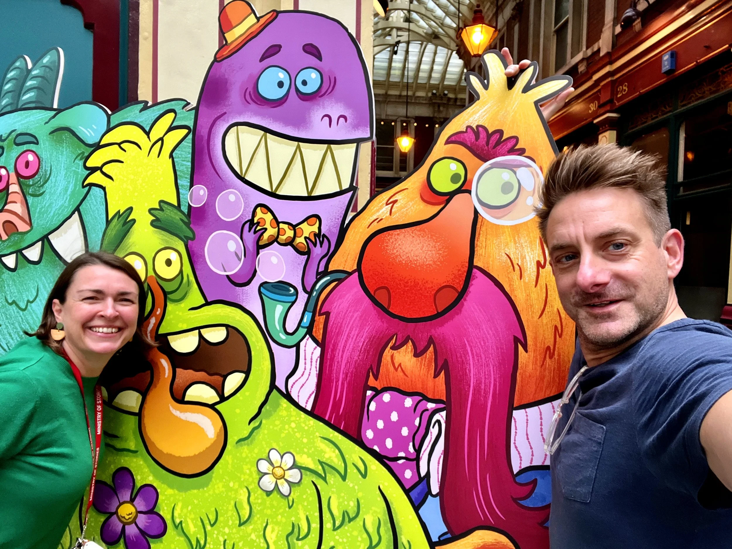 A smiling woman wearing a green hoodie and a man with light brown hair wearing a dark shirt take a selfie in front of colorful cartoon monster cutouts inside a market or arcade with glass ceiling and hanging lights.