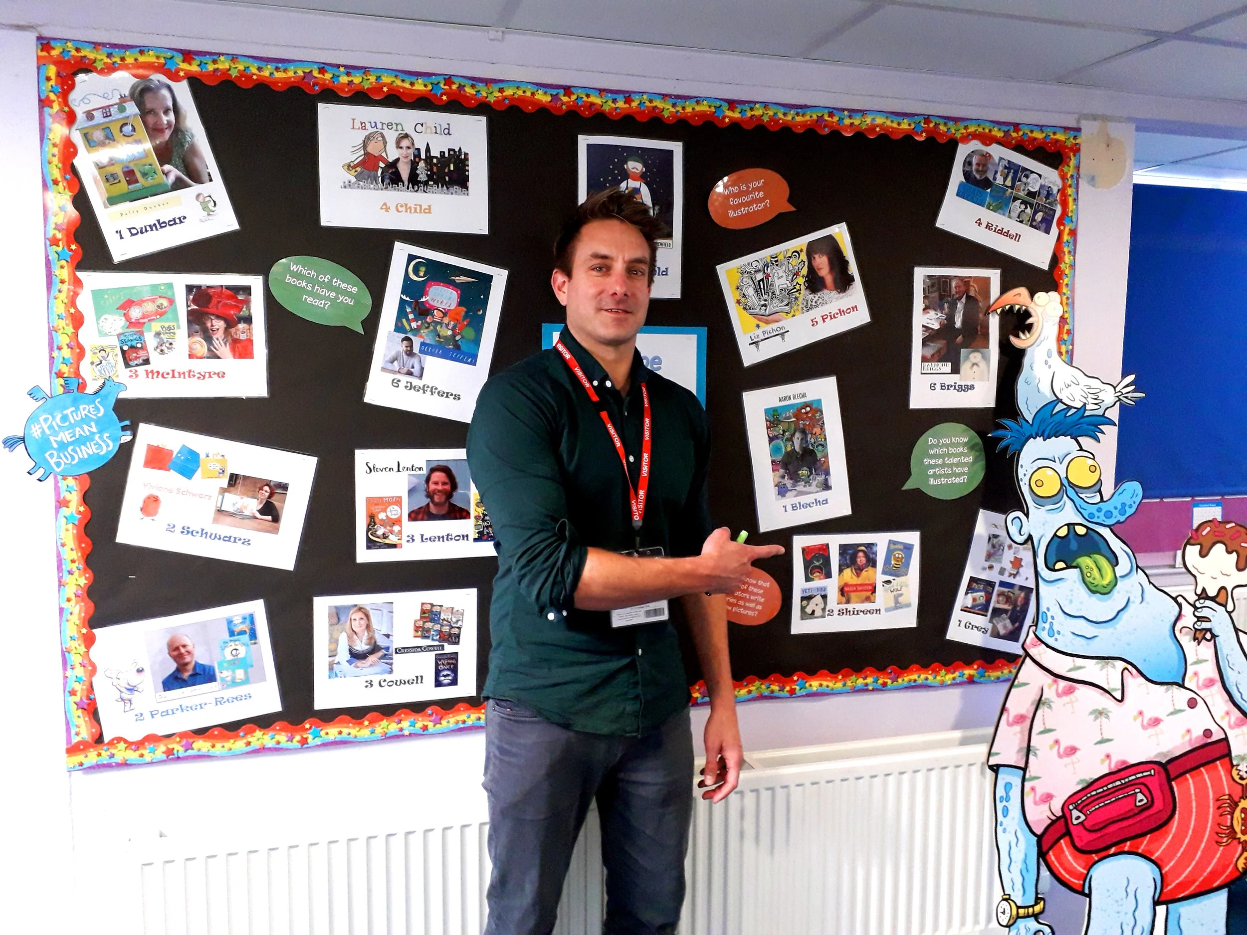 A man standing in front of a colorful classroom display board featuring photographs and names of various authors, along with prompts about books and illustrators. To his right, there is a large cartoon cutout of a girl with glasses and a white dress with pink flamingos, and a cartoon dinosaur with a big mouth, glasses, and wearing a dress.
