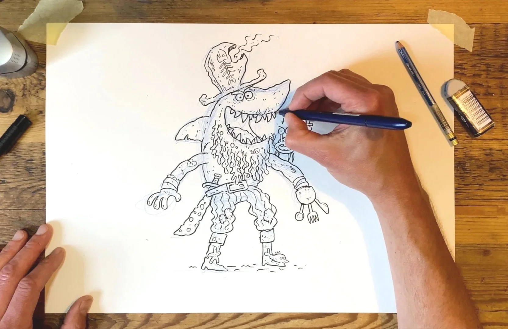 Person drawing a cartoon pirate monster with a beard, hookhand, eye patch, and pirate accessories on a large sheet of paper on a wooden table.