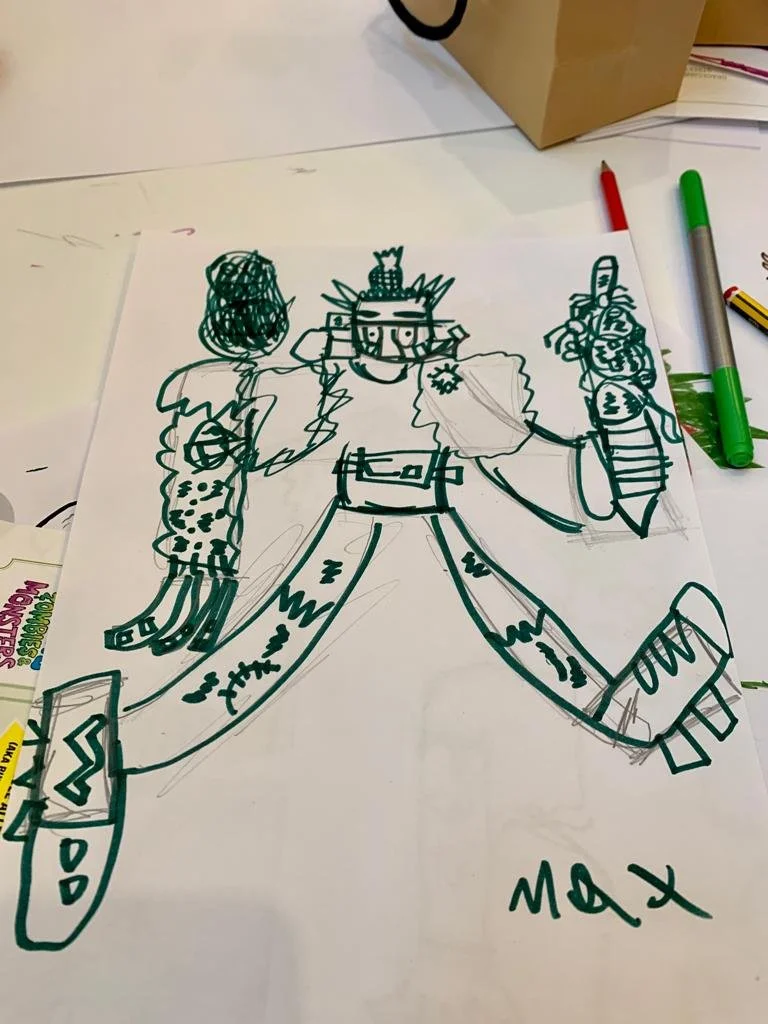 A hand-drawn cartoon of a Viking with a beard, wearing a helmet with horns, holding a sword in one hand and a shield in the other. The drawing is on a piece of white paper on a table with markers nearby, and the name "Max" is written at the bottom.