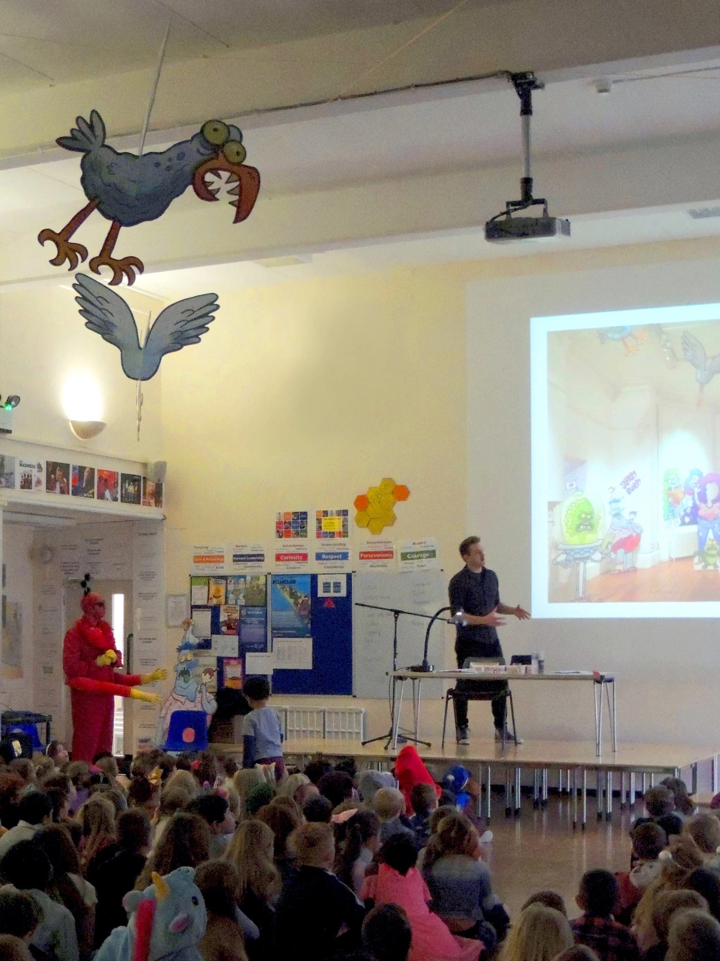 A classroom or auditorium filled with children sitting on the floor watching a presentation or performance on stage. On stage, a man is speaking, and some children are dressed in costumes, including a person in a pink costume and another in a unicorn costume. Behind him, a large screen displays a colorful cartoon image. There is a hanging decoration of a cartoon bird with large eyes and an open beak.