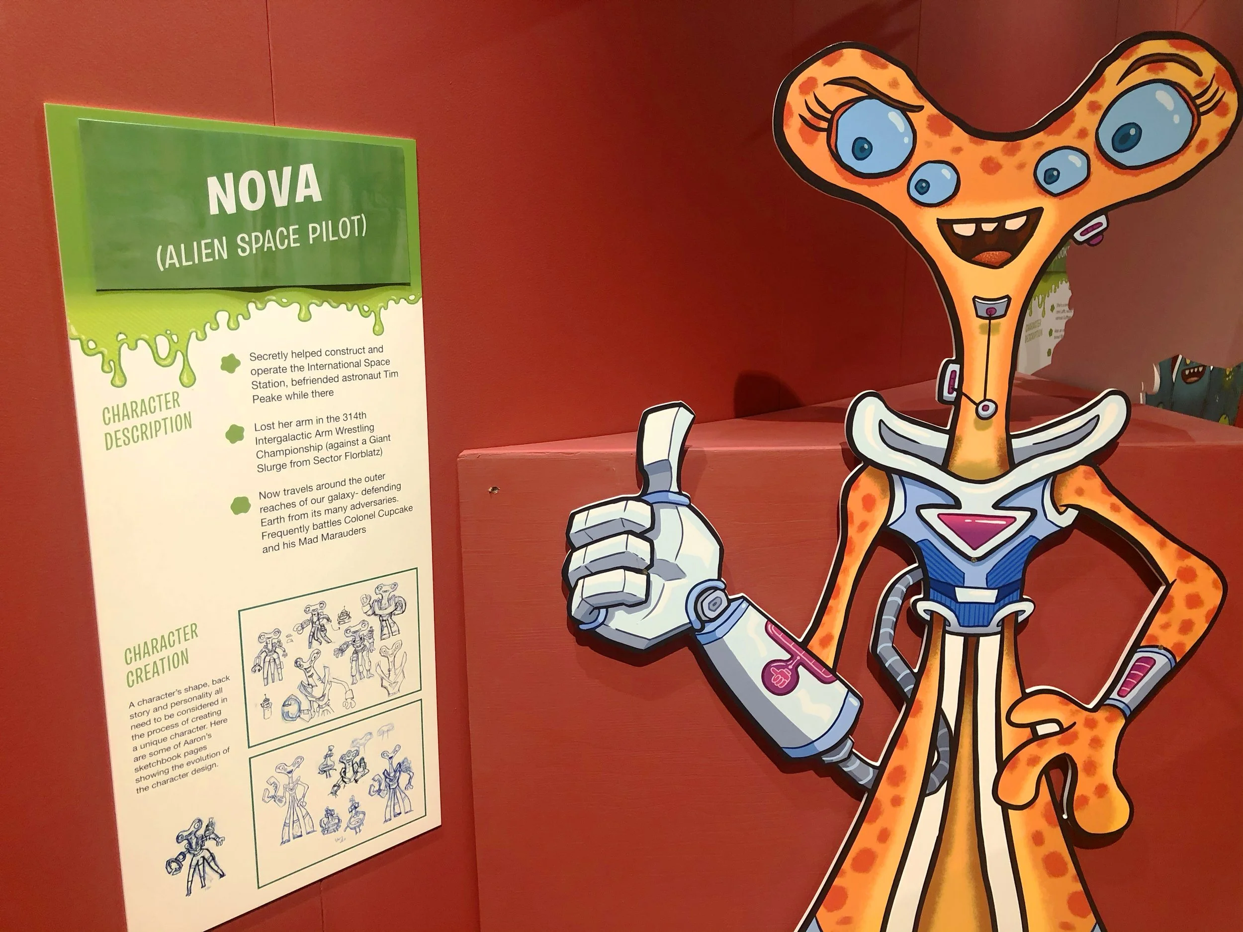 Colorful display with a cartoon character of a giraffe with multiple blue eyes, wearing a superhero outfit, giving a thumbs up, standing next to an informational poster about Nova, an alien space pilot.