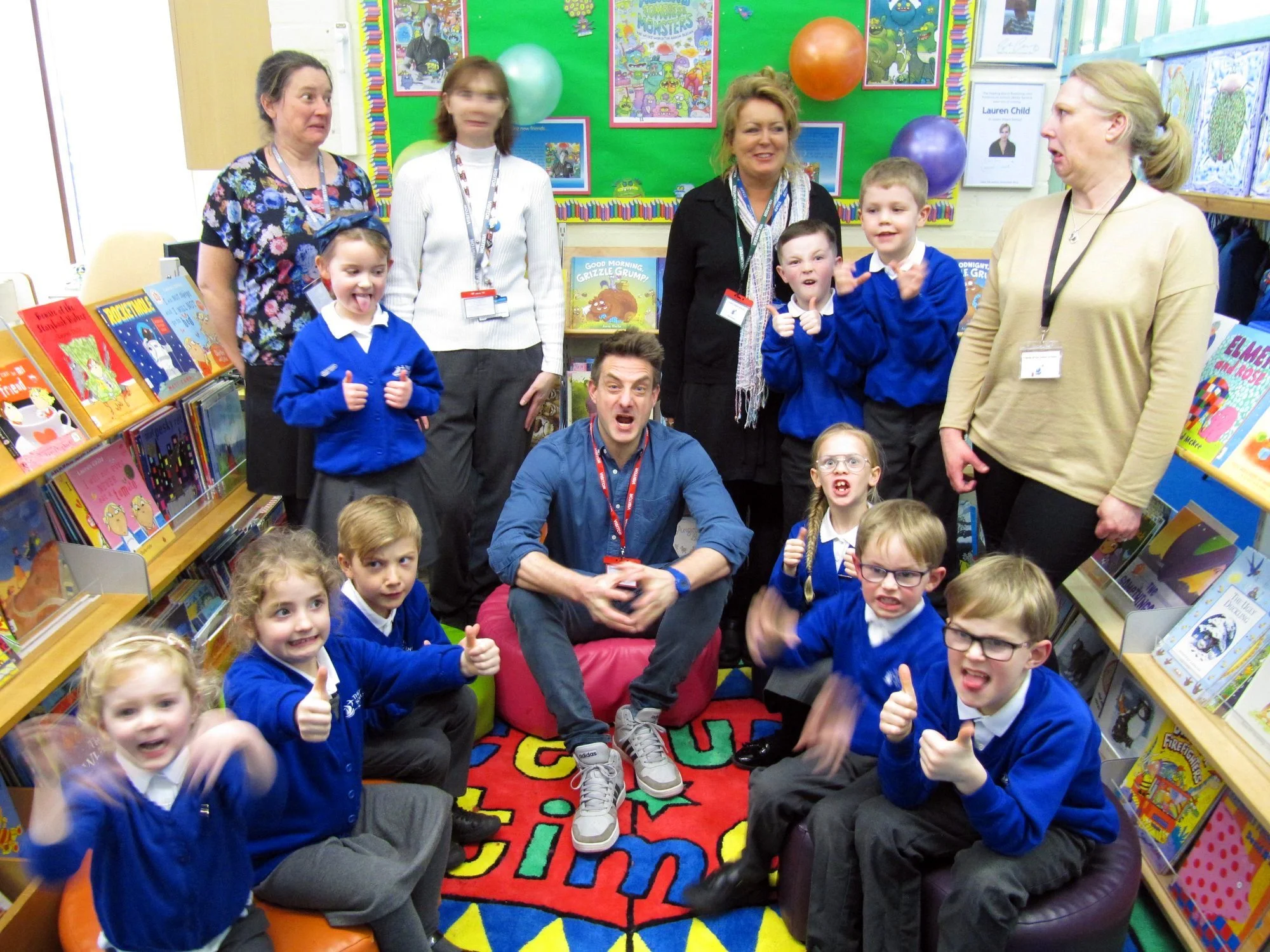 Children and adults in a colorful library or bookstore, with bookshelves filled with children's books, some of the kids are sitting on a multicolored rug while others stand around, all smiling and making funny faces.