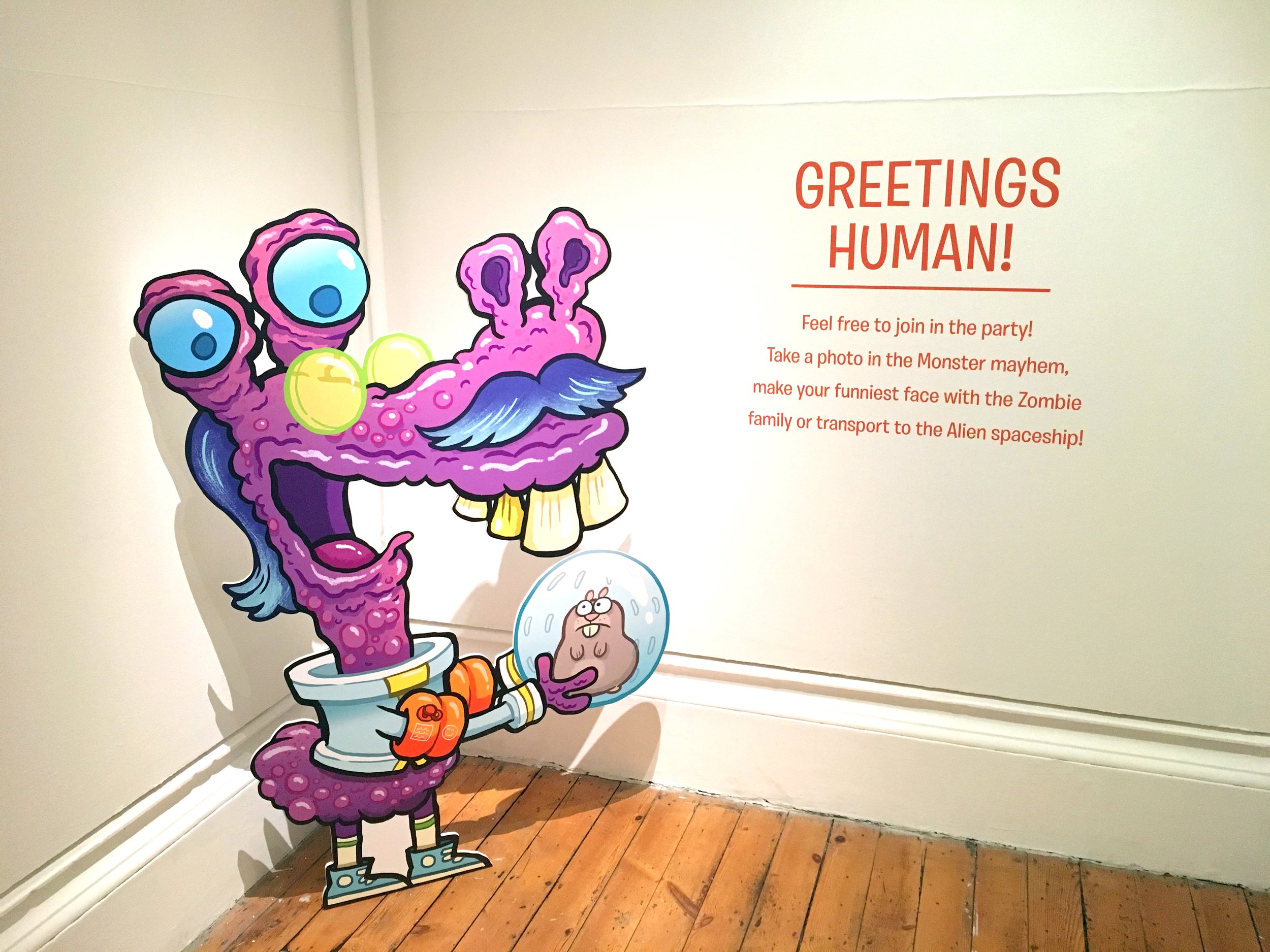 Colorful cardboard cutout of a purple monster with big blue eyes, a mustache, and a wide open mouth, sitting on a toilet with a roll of toilet paper, while wearing star-shaped sunglasses, next to a small character with glasses encased in a clear ball.