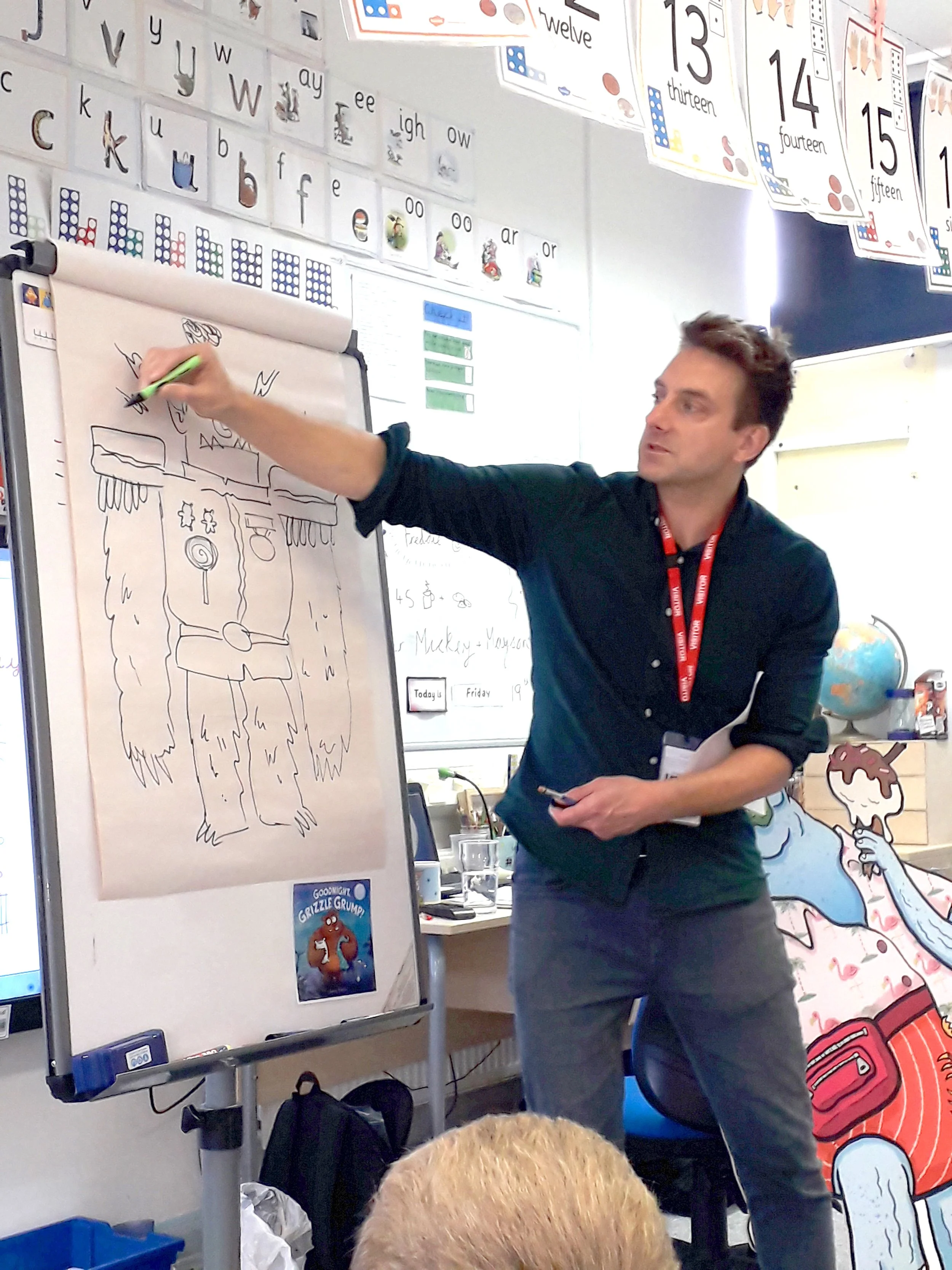 A man is teaching a classroom lesson using a whiteboard with a large drawing of a creature with fur and claws. The teacher is pointing at the drawing with a green marker. The classroom wall behind him displays alphabet and number posters, including numbers thirteen to fifteen.