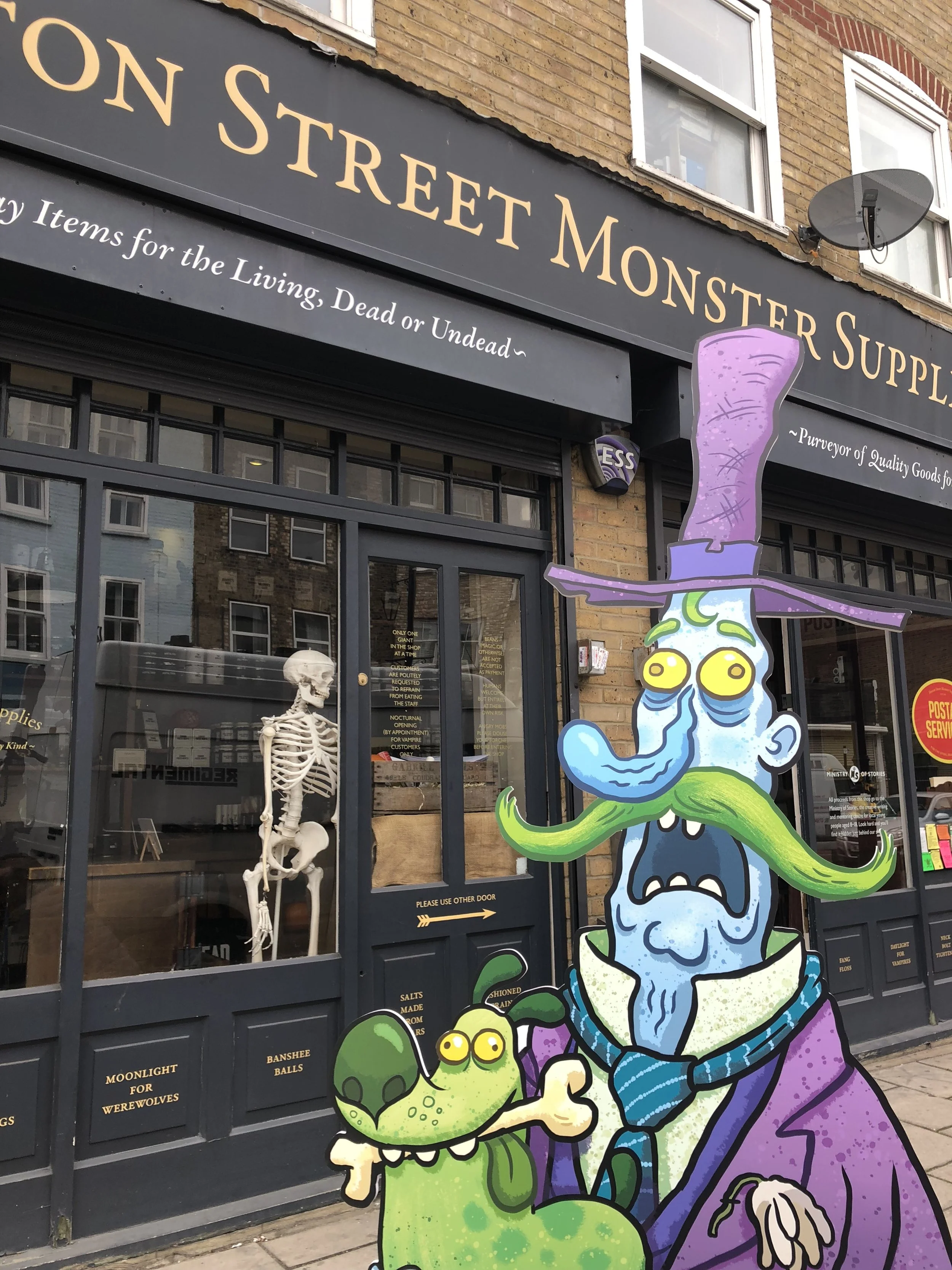 Colorful cartoon illustration of a witch with long purple hair, green tongue, and yellow eyes, holding a green monster with yellow eyes and a bone in its mouth, standing outside a store named 'On Street Monster Supplies' with a skull skeleton display behind the glass door.
