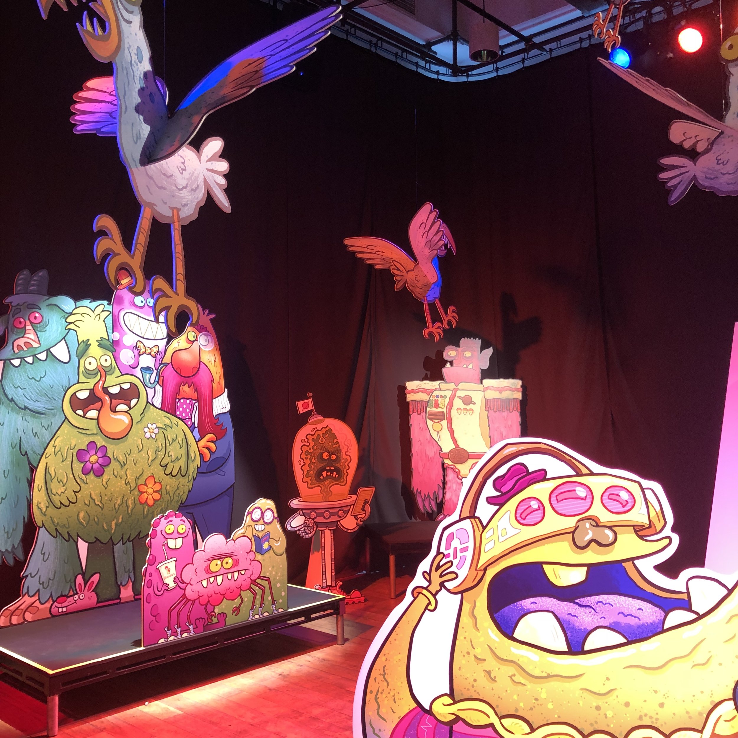 Colorful cartoon-style display featuring various whimsical monsters, animals, and a large pizza with toppings, illuminated by neon and stage lighting.
