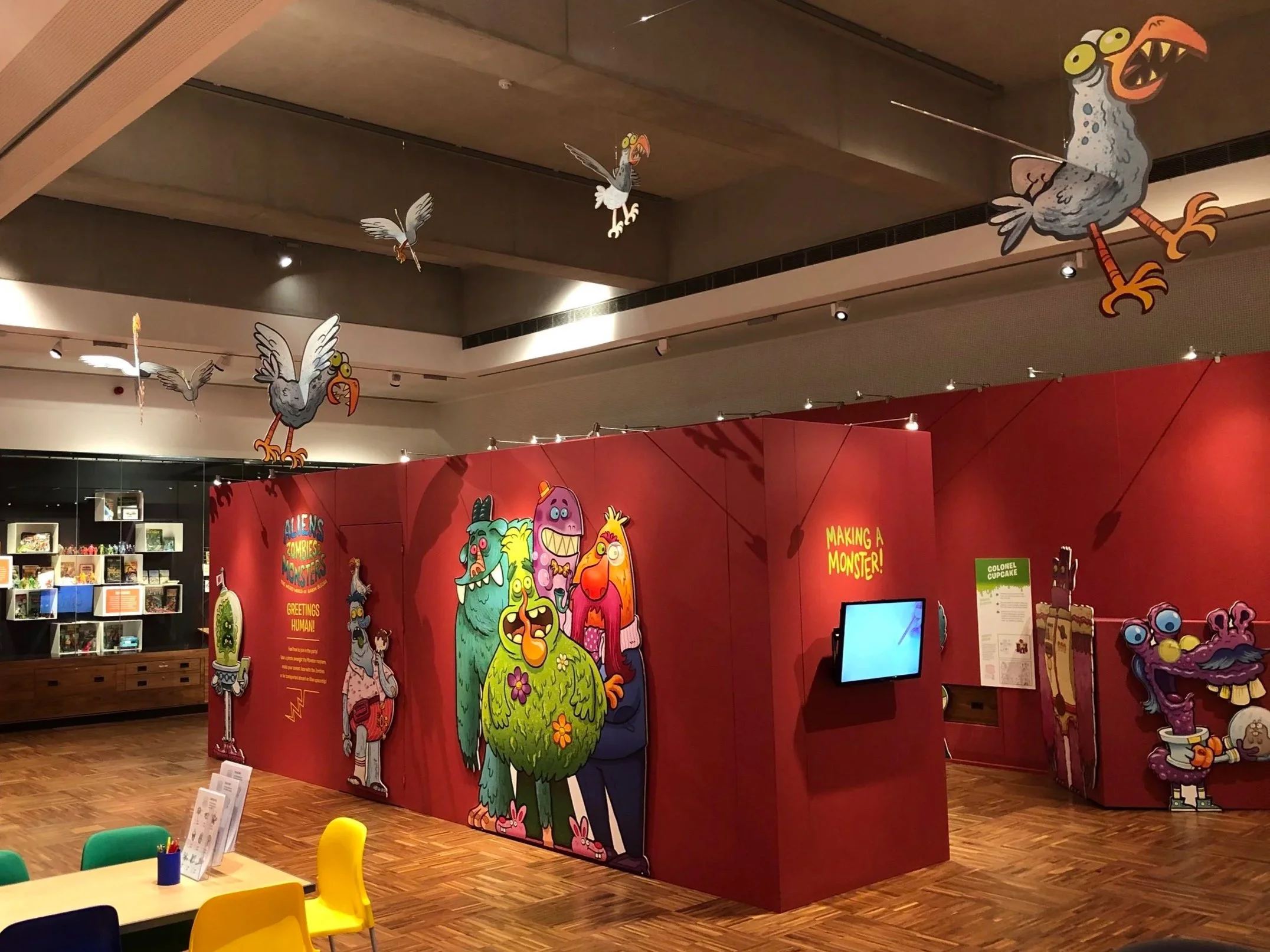Colorful display in a museum or exhibition featuring cartoon monsters, with a red background and various monster characters including a large green monster with pink flowers, a purple monster with one eye, and other quirky creatures. Hanging cartoon bird figures are suspended from the ceiling.