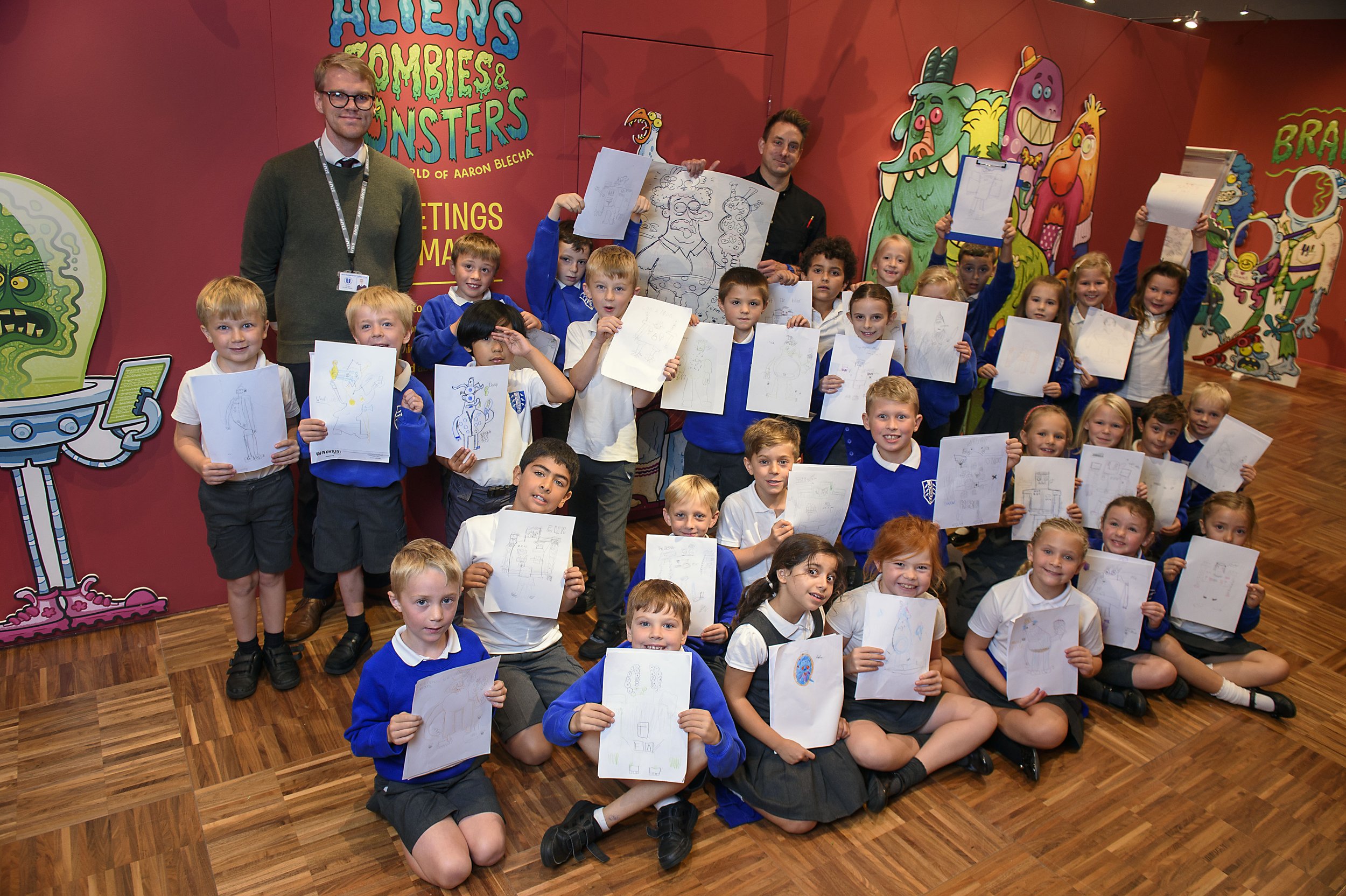 A group of children holding up drawings in front of a colorful cartoon backdrop, with two adults standing behind them at an event related to aliens, zombies, and monsters.