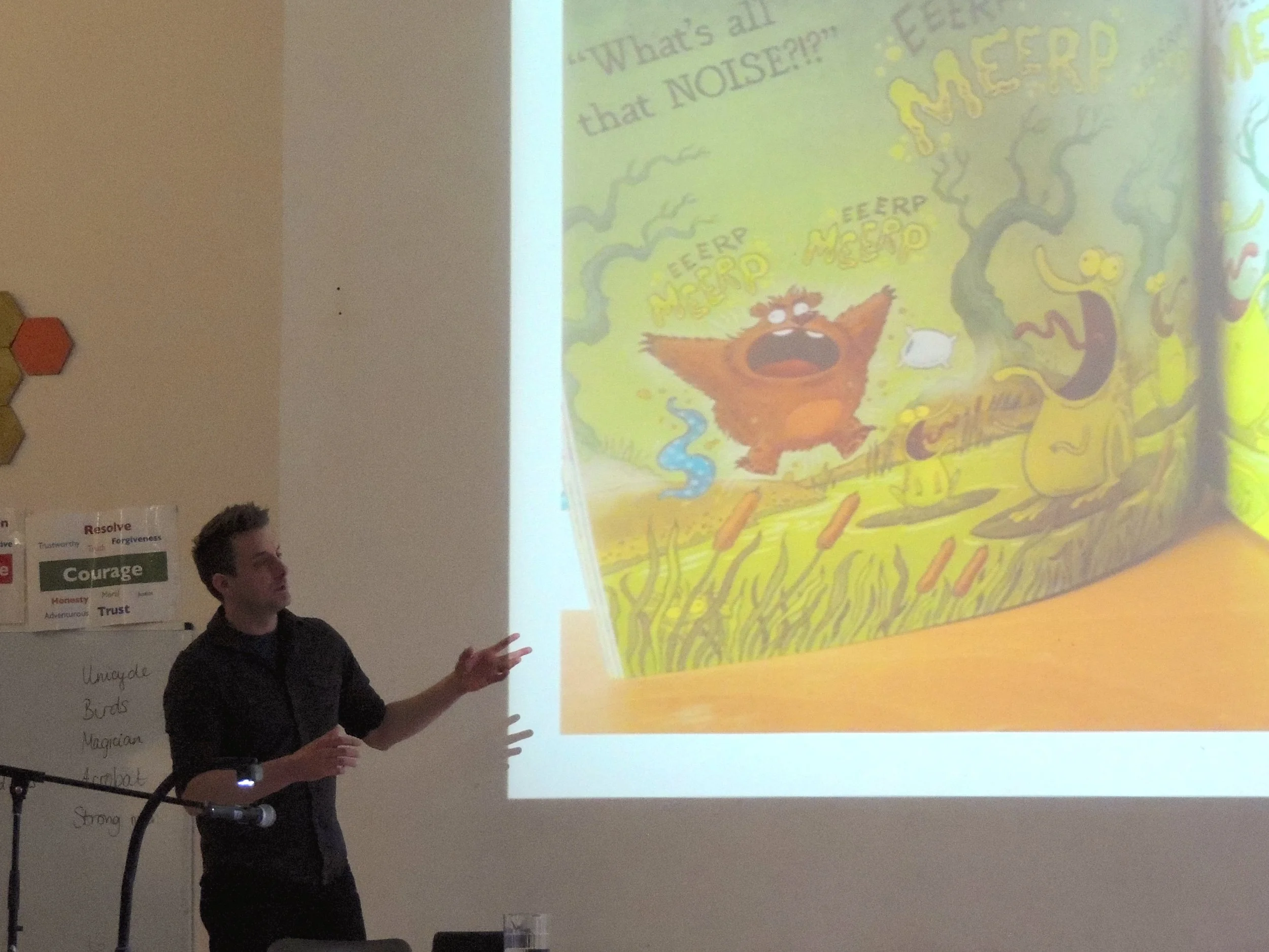 A person giving a presentation in front of a projection screen displaying a colorful cartoon illustration of a tree with a creature and a bird, with text "What’s all... that NOISE?!?" at the top.