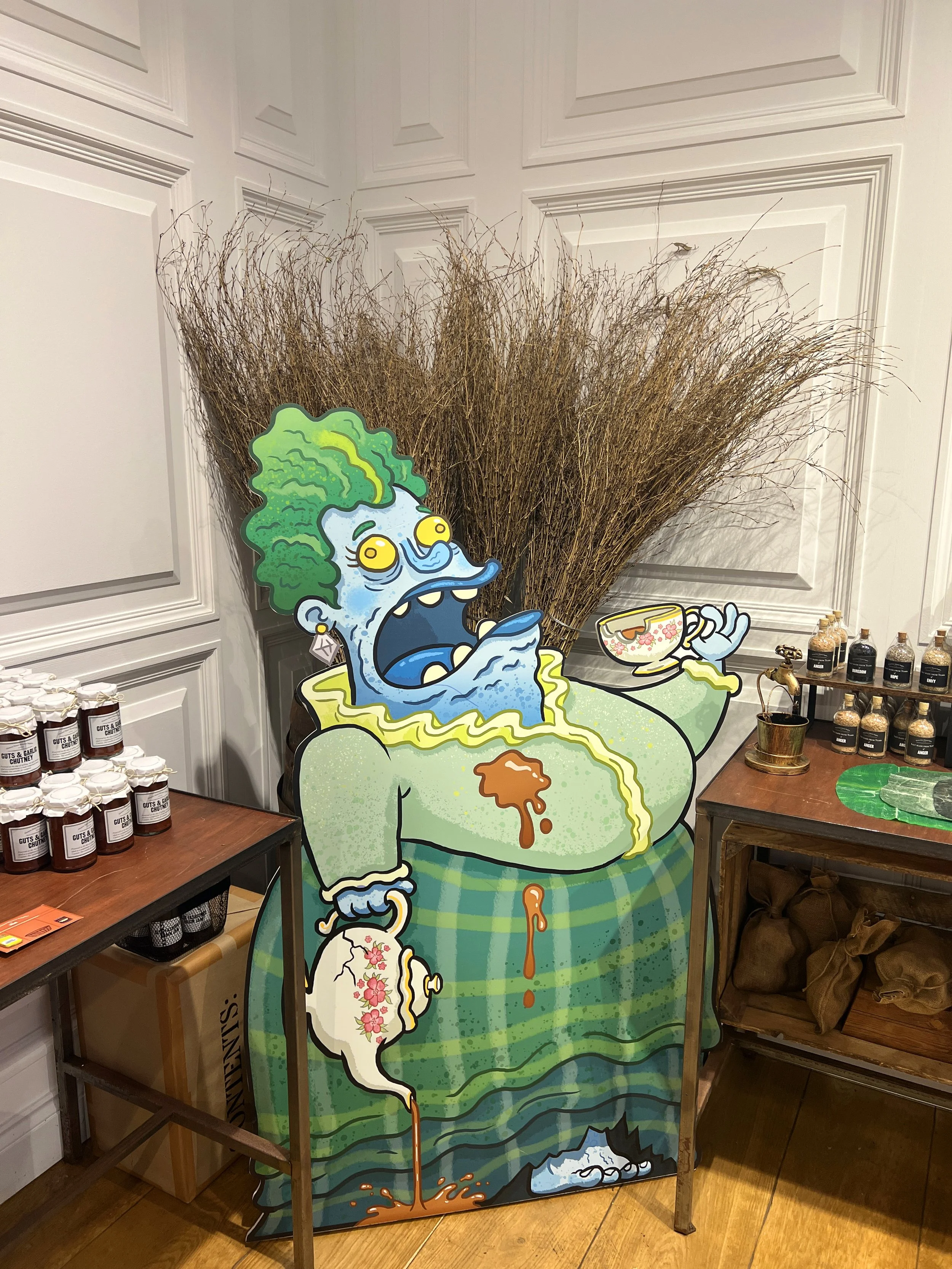 A large, colorful cartoon cutout of a person eating soup, with exaggerated features and green hair, set against a white paneled wall. There are tables with jars and bottles nearby.