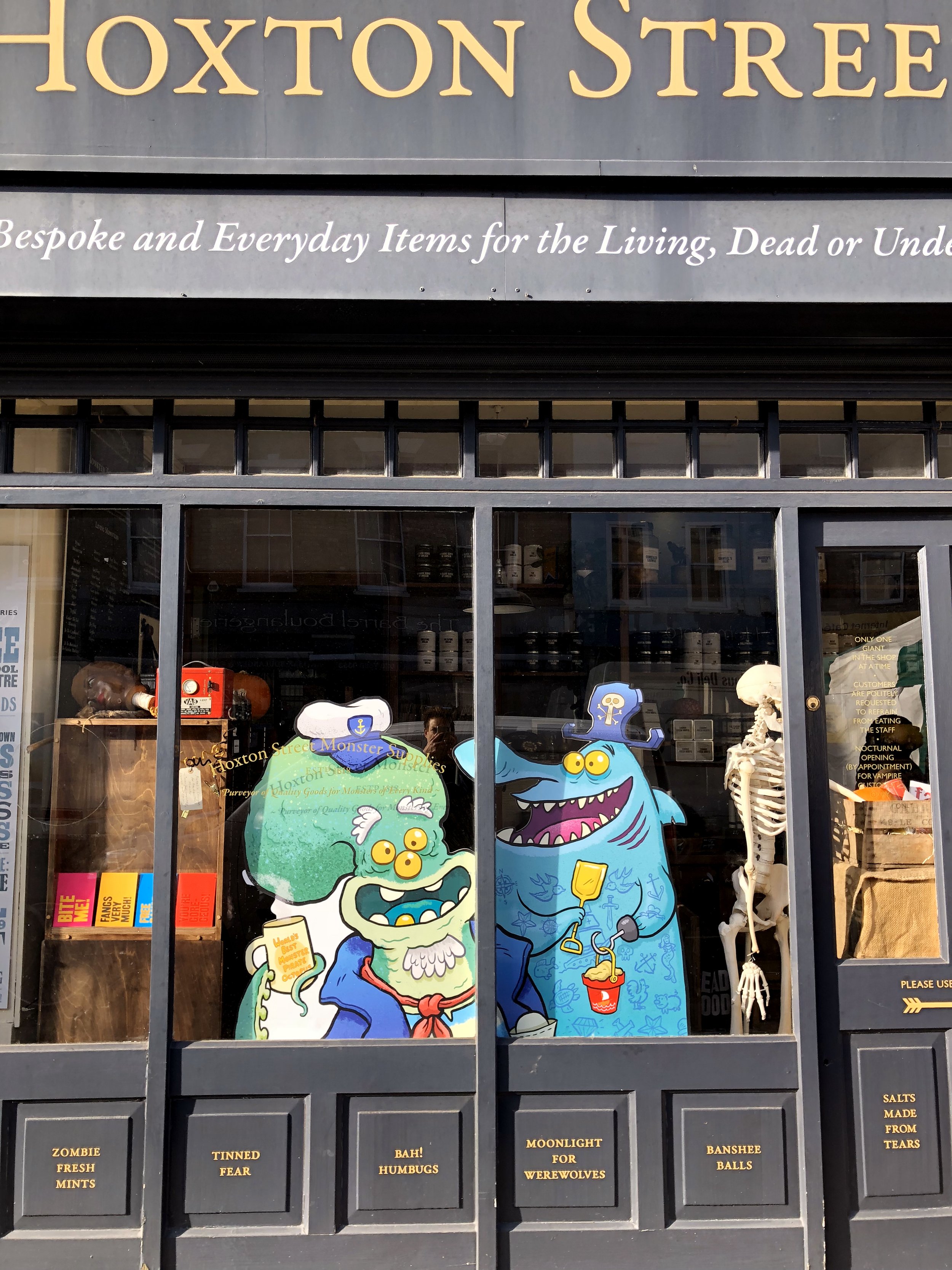 Store window with Halloween-themed cartoon character cutouts, including a green monster with three eyes and a blue monster with a skeleton, with Halloween decorations and items visible inside.