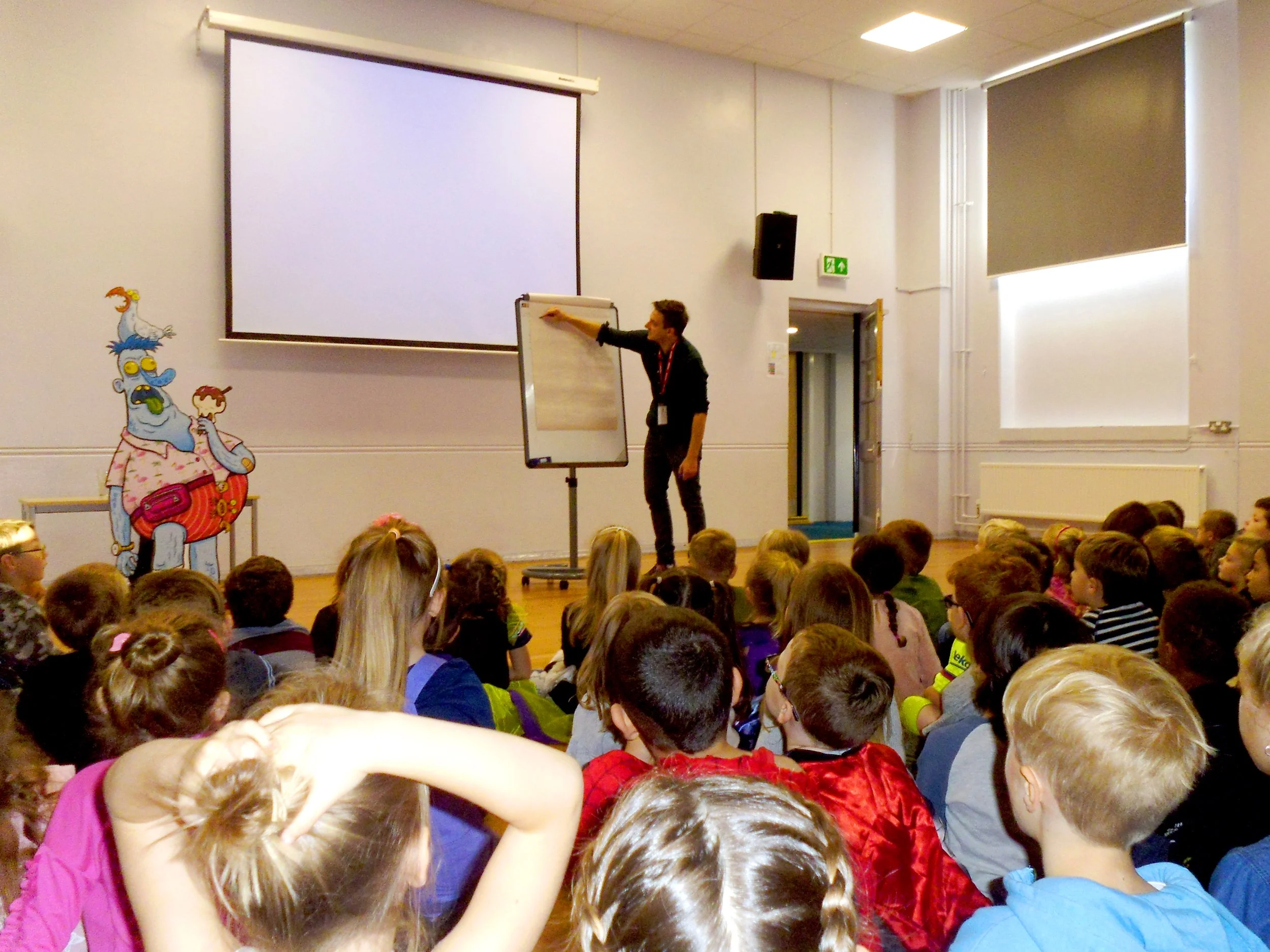 A man giving a presentation in front of a large audience of children in a classroom or auditorium. The children are seated on the floor, facing the speaker. There is a whiteboard, a large projection screen, and colorful cartoon wall decals.