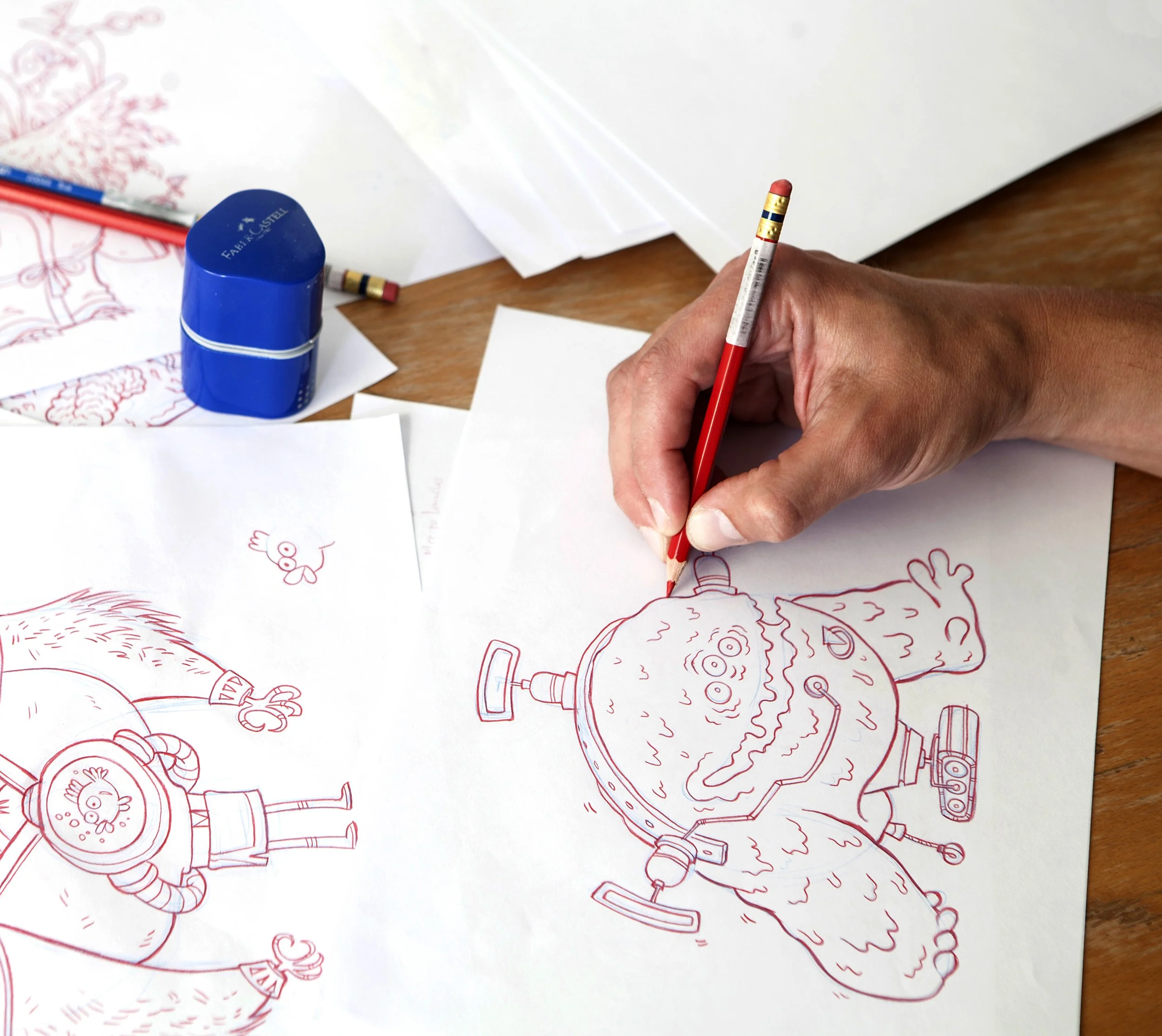 A hand holding a red colored pencil drawing on a sheet of paper with red outline sketches of various cartoonish characters, including a robot, a snowman, and a person in a helmet, on a wooden surface. There are some sheets of paper and a blue glue stick nearby.