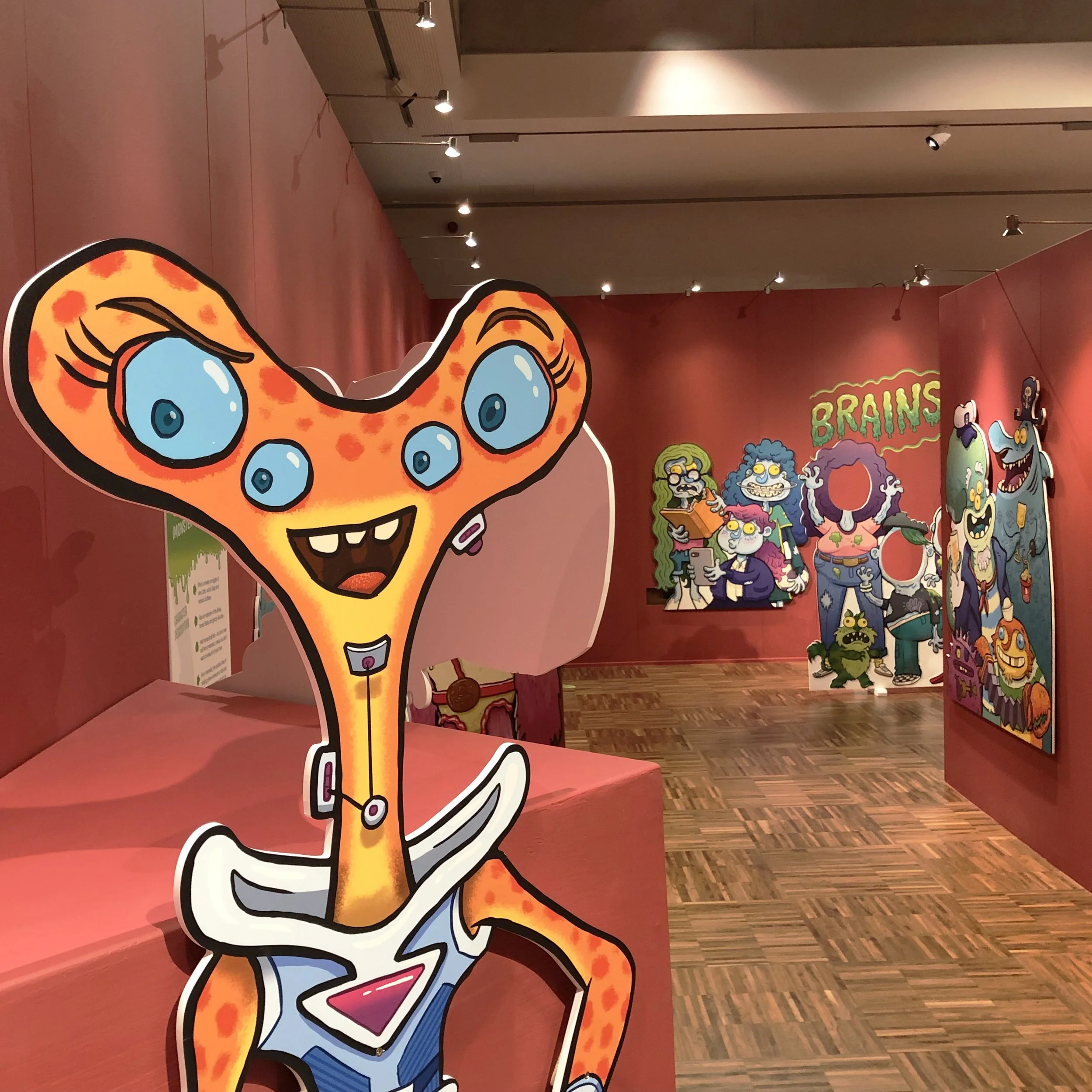 Colorful cartoon-style characters at an exhibit with a pink background, featuring a large cartoon leopard with blue eyes and a big smile, and various other monster characters in the background.