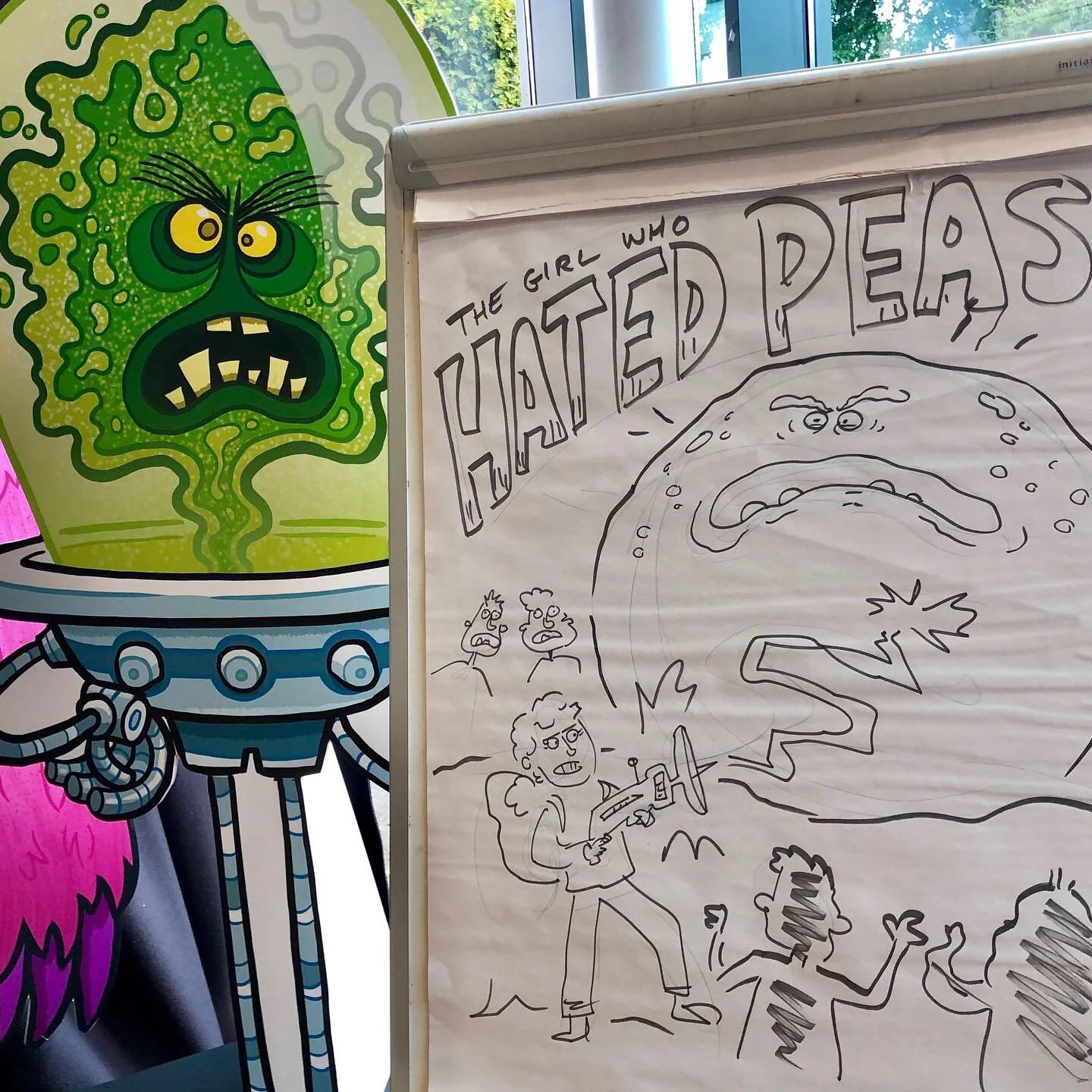A black and white drawing on a large sheet of paper depicts a scene with a giant monster and a person with a gun, fighting or battling. The scene is labeled "The Girl Who Created Peas" at the top. There are smaller characters around the scene showing fear and action.