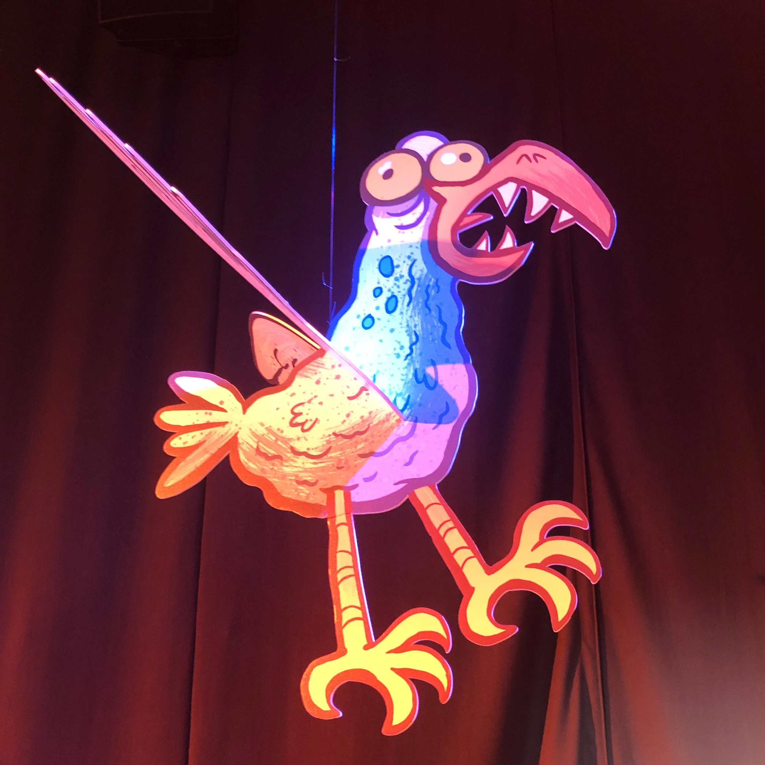 Brightly colored neon sign of a cartoon dinosaur with an open mouth, large eyes, and a floral design on its body, set against a dark background.
