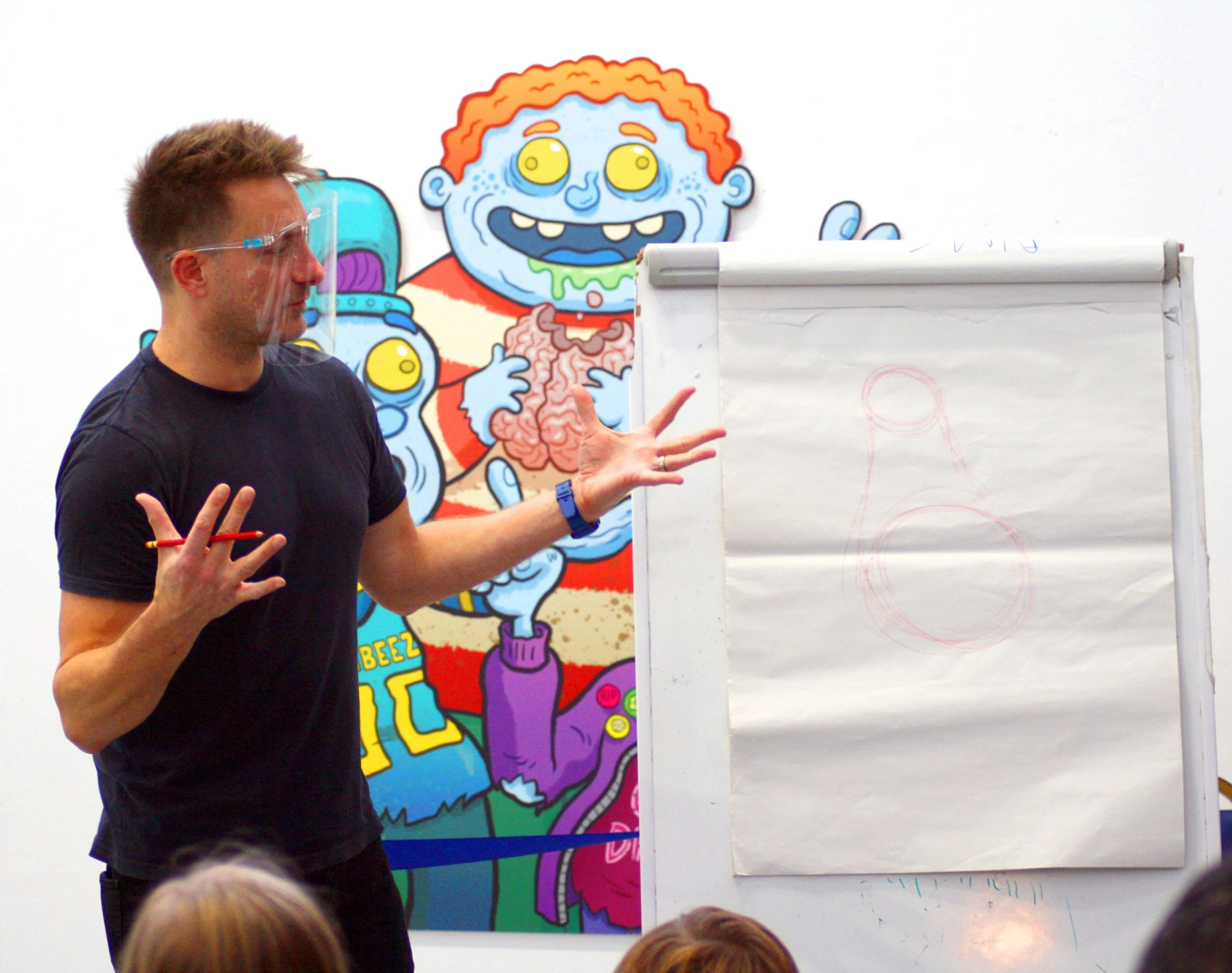 A man is giving a presentation in front of a colorful cartoon zombie illustration on a white wall, with children in the foreground listening.