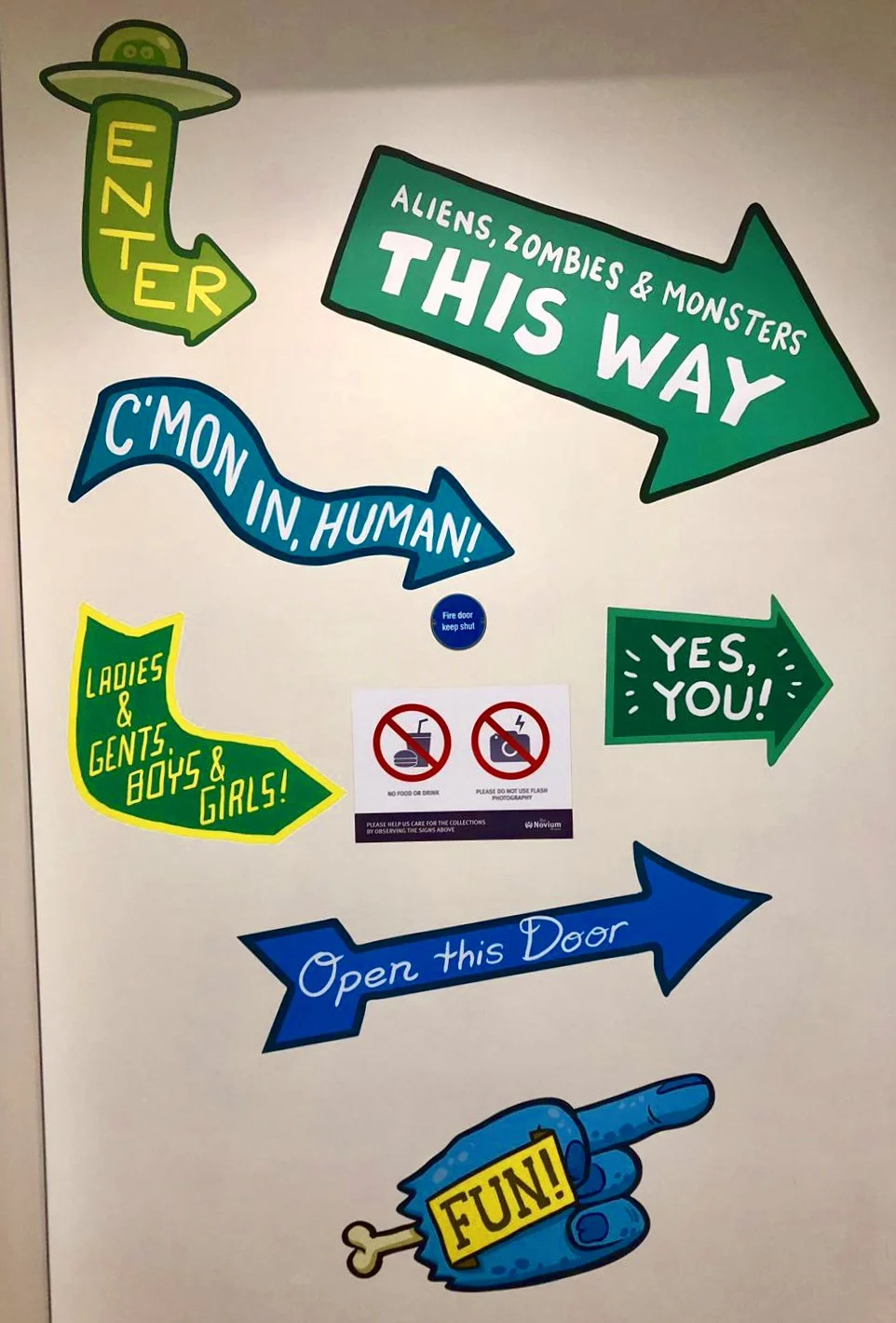 Colorful signs and arrows directing people for a themed event, with messages like ‘Aliens, Zombies & Monsters This Way’, ‘C'mon in, Human!’, ‘Yes, You!’, ‘Ladies & Gents, Boys & Girls!’, ‘Open this Door’, and a pointing hand with a sign reading ‘FUN!’. There are also two prohibition signs for food or drink and flash photography, and a small blue sign saying ‘Fire door keep shut’.