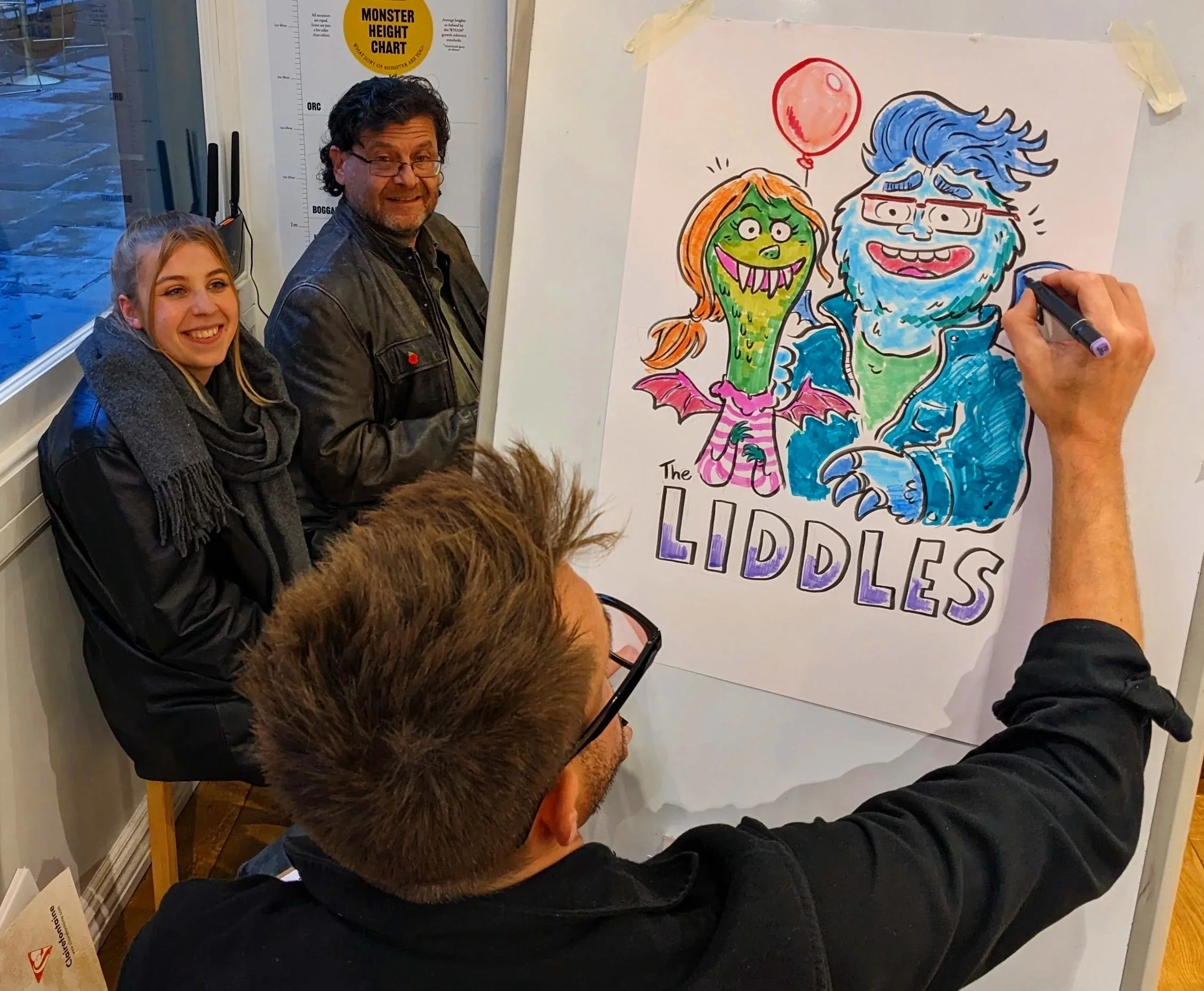 People gathered around a drawing of a colorful cartoon monster and a smiling blue-haired man with glasses on a whiteboard. The drawing includes a green monster with orange hair, a red balloon, and the words 'The LIDDLES' written at the bottom.