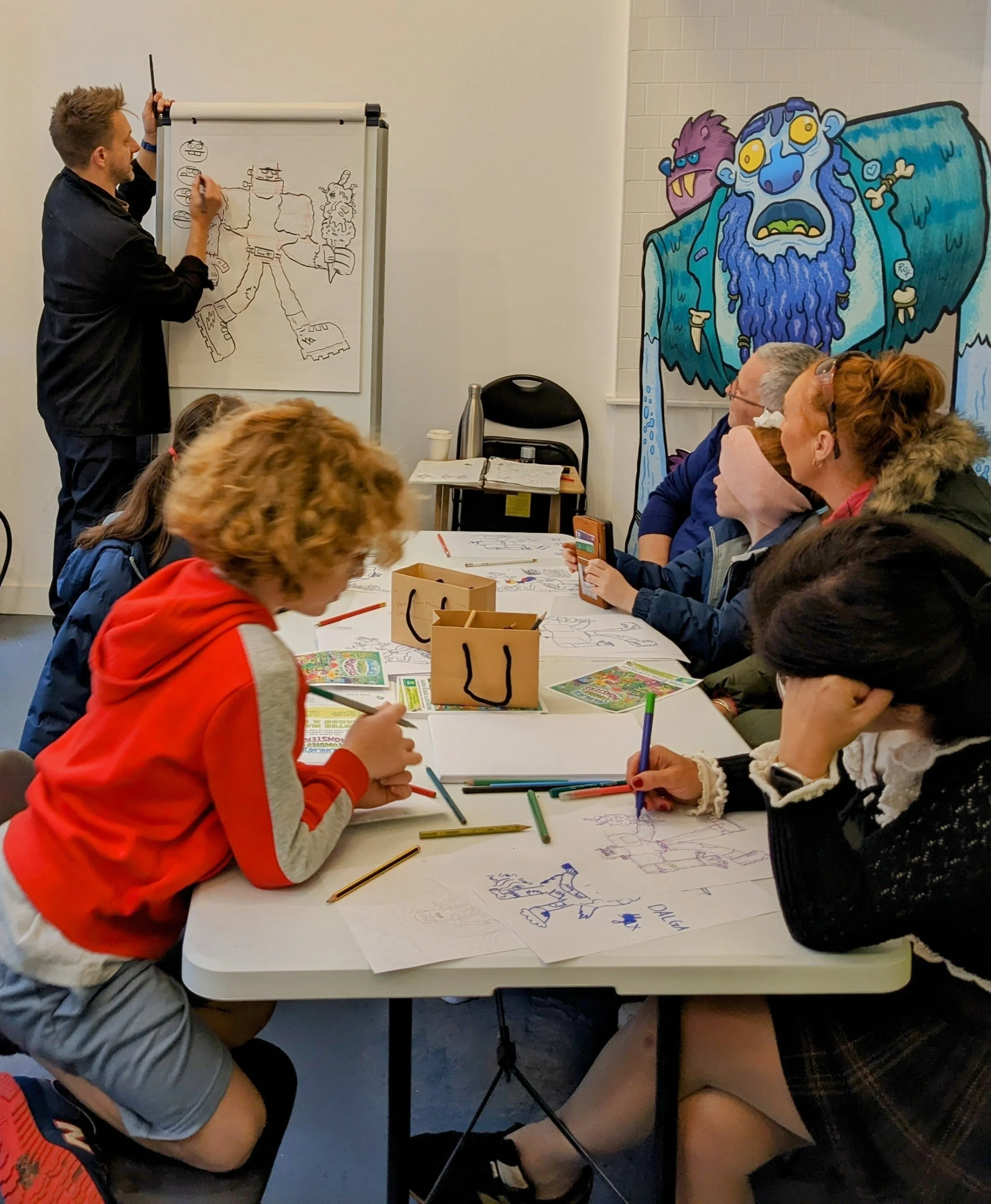 A group of children and adults sitting around a table, coloring and drawing on large sheets of paper. An instructor is drawing on a whiteboard in the background, and there is colorful wall art depicting cartoon monsters.