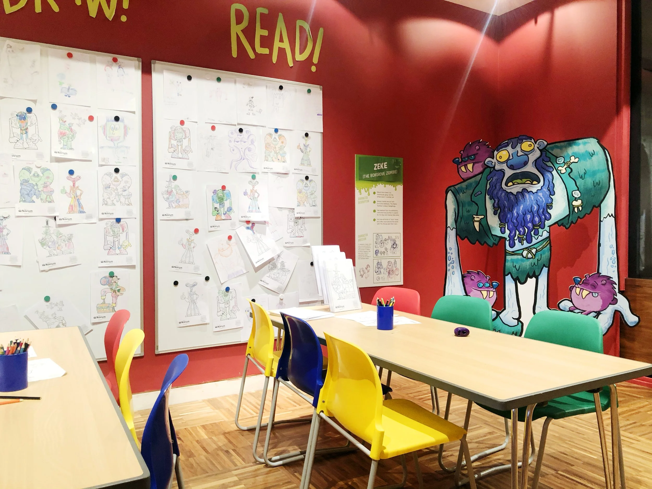 Colorful classroom with tables and chairs, decorated walls with drawings and a large mural of a monster, and educational posters.