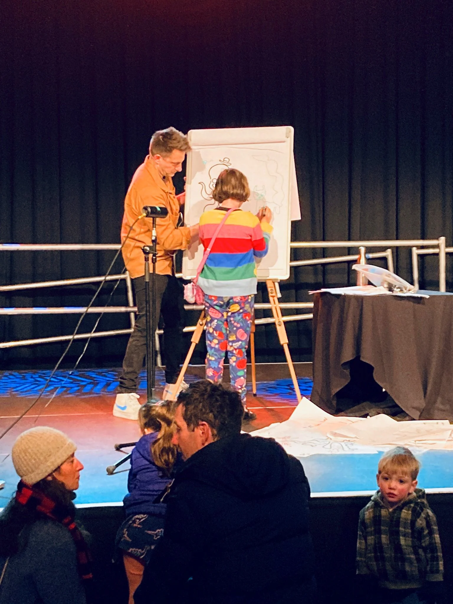 Children and adults seated on the floor watching a performer drawing on a whiteboard during a show on a stage with black curtains.