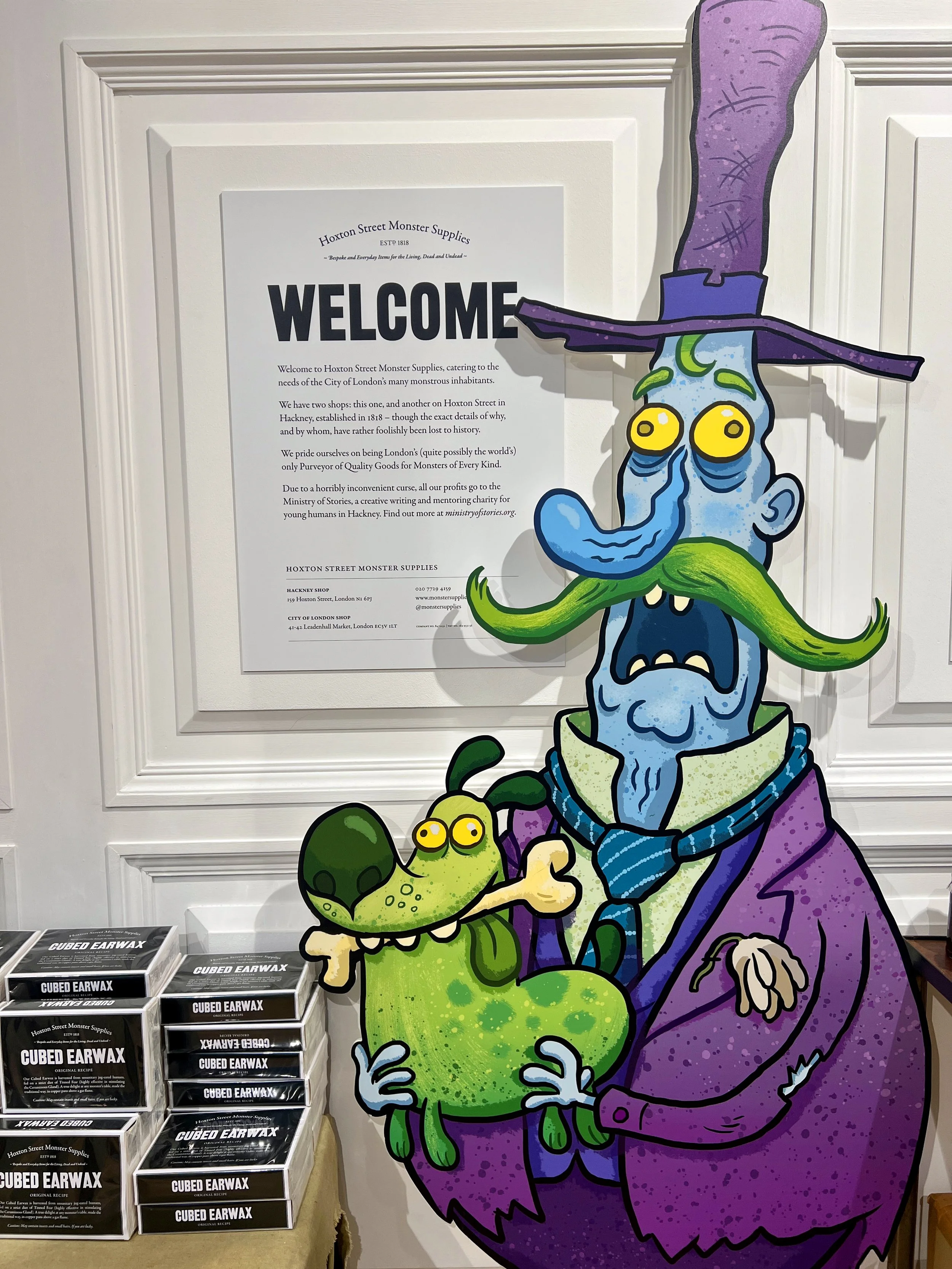 Cartoon illustration of a witch with purple hair and green beard holding a small green creature with yellow eyes, standing in front of a sign for Hoxton Street Monster Supplies.